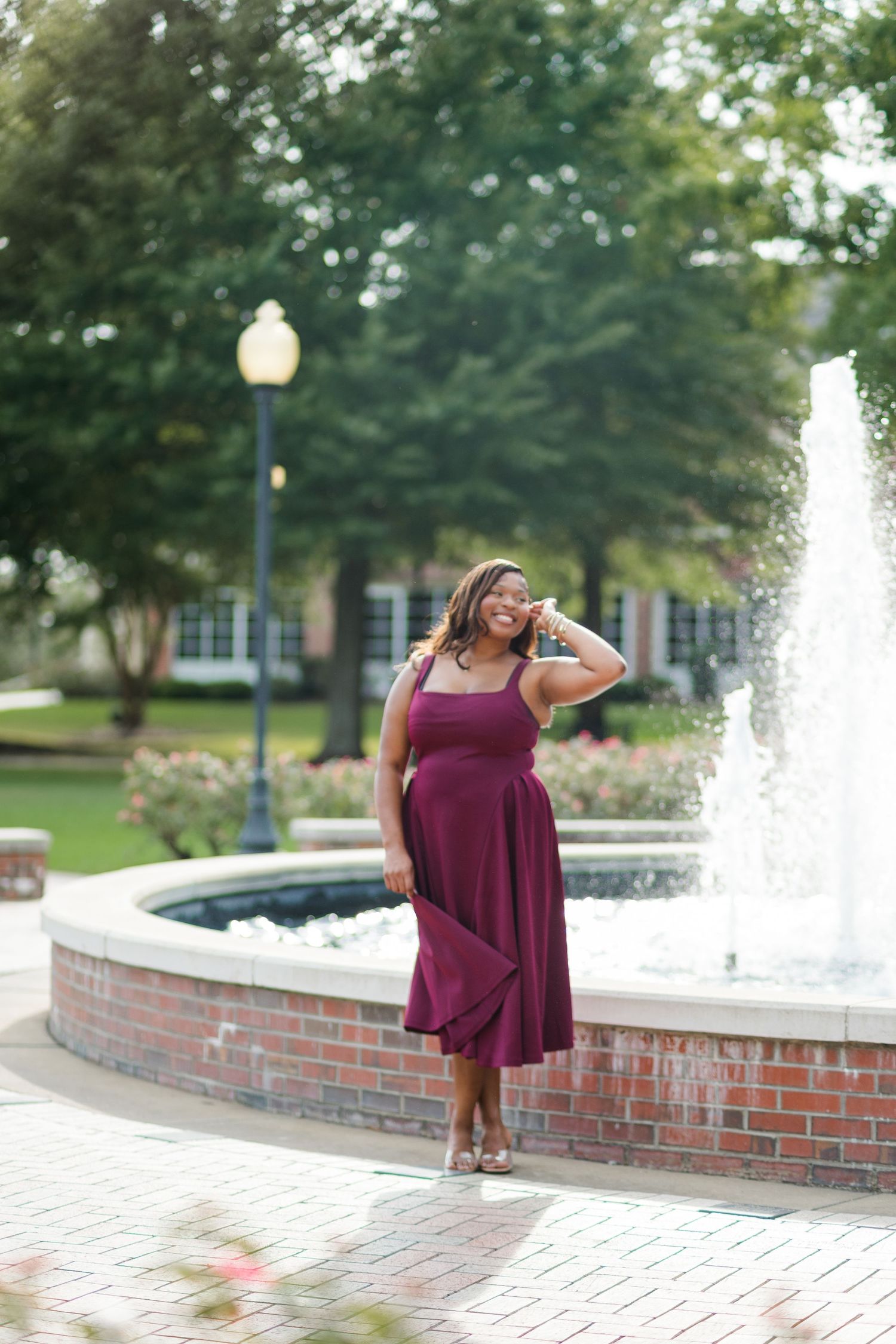 Woman in burgundy midi dress posing by brick fountain in park setting with green trees and lamppost.