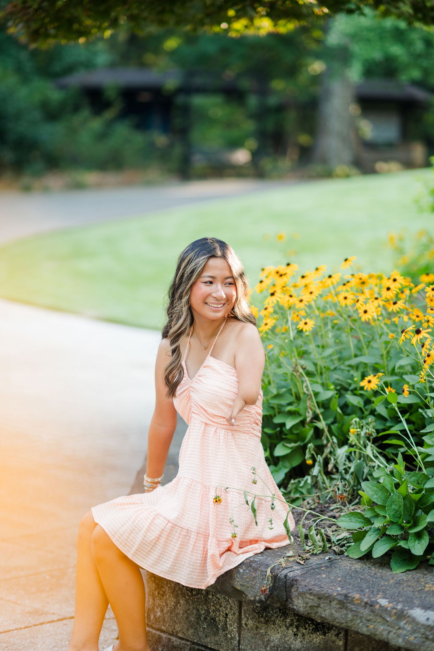 Smiling young woman in peach dress perched on stone wall near black-eyed Susans.