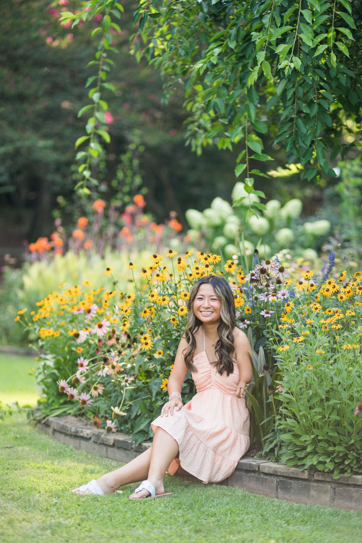Young woman in pink dress sitting by colorful garden flowers, smiling outdoors.