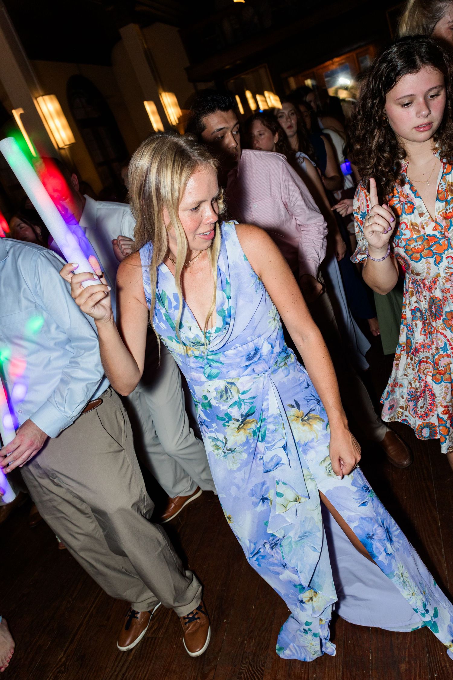 Guests dancing at a wedding reception, woman in blue floral dress holding a light-up foam stick on the dance floor.