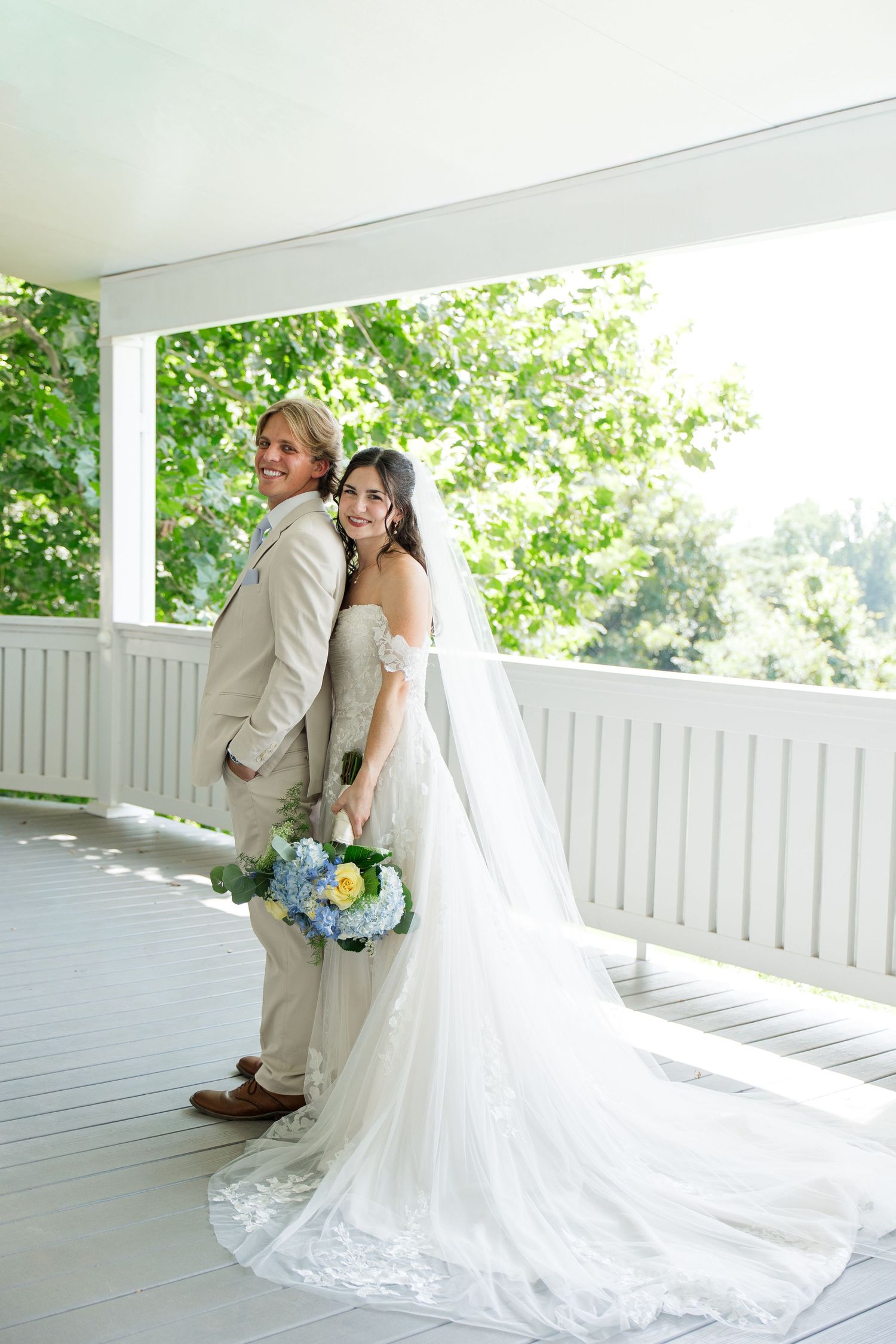 Bride and groom pose together on white porch with lush green trees, bride holding blue and yellow bouquet.