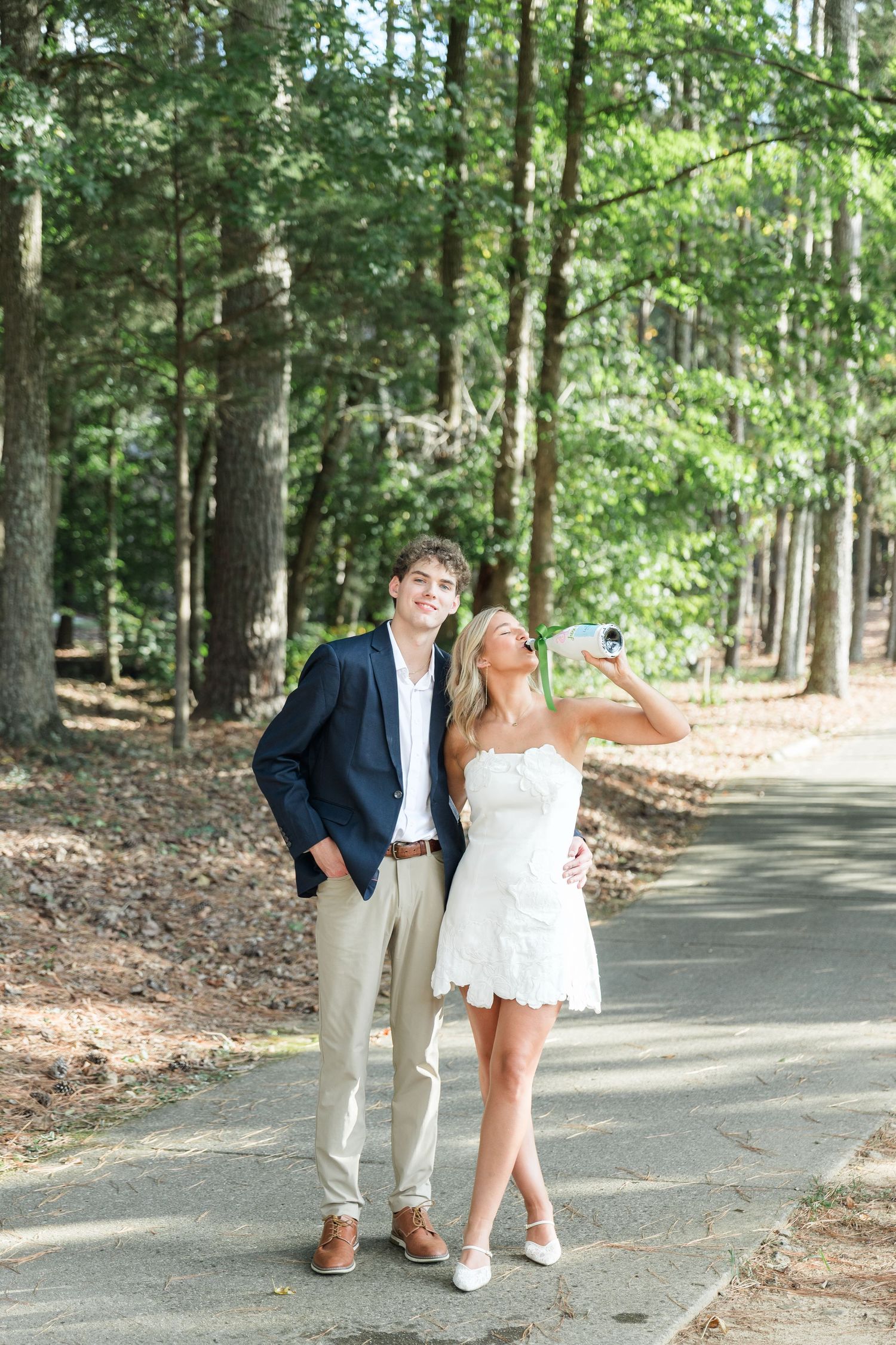Smiling couple posing on a shaded path, woman in white dress taking a sip from a bottle.