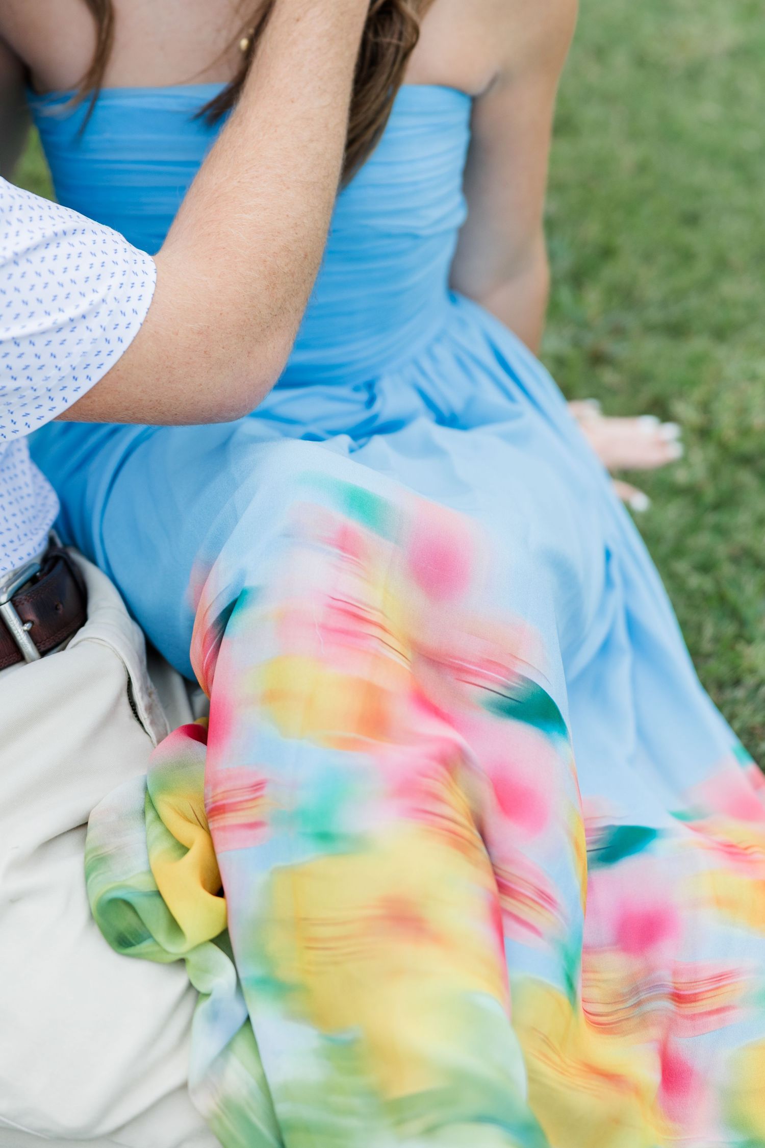 Woman in a blue strapless dress with colorful watercolor skirt sits on grass while a man in a white shirt rests his arm on her shoulder.