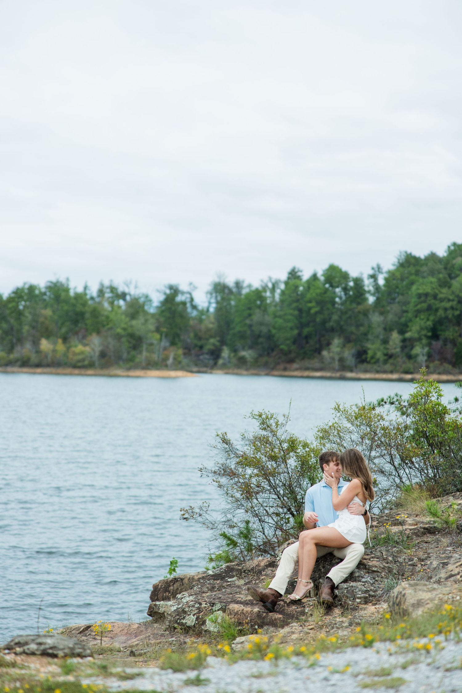 Couple sits close together on a rocky outcrop beside a quiet lake with wildflowers and forested shoreline in the background.