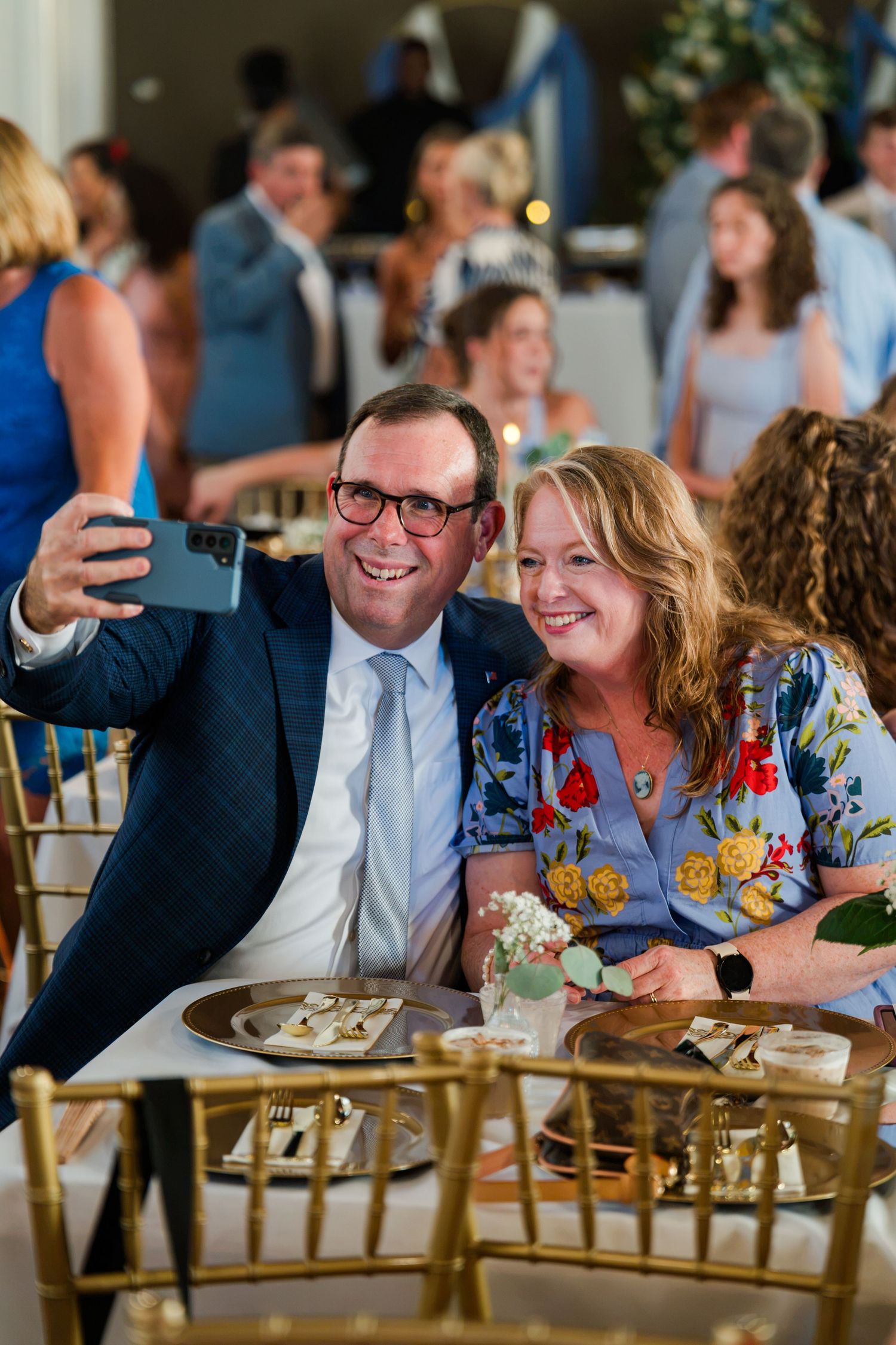 A man in a navy suit and a woman in a floral dress take a smiling selfie at an elegant wedding reception.
