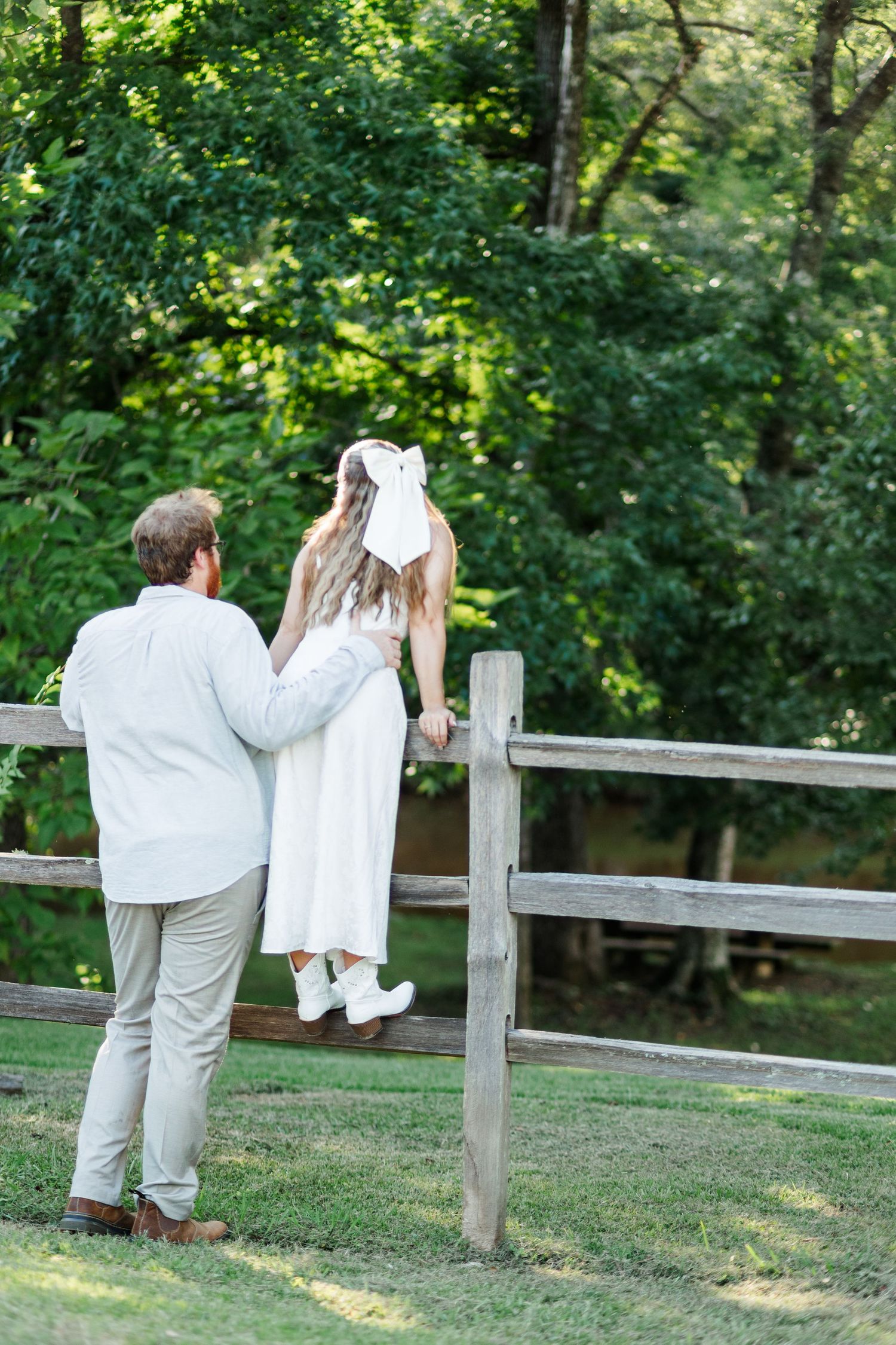 A couple in white attire stands by a rustic wooden fence surrounded by lush green trees in a serene outdoor setting.