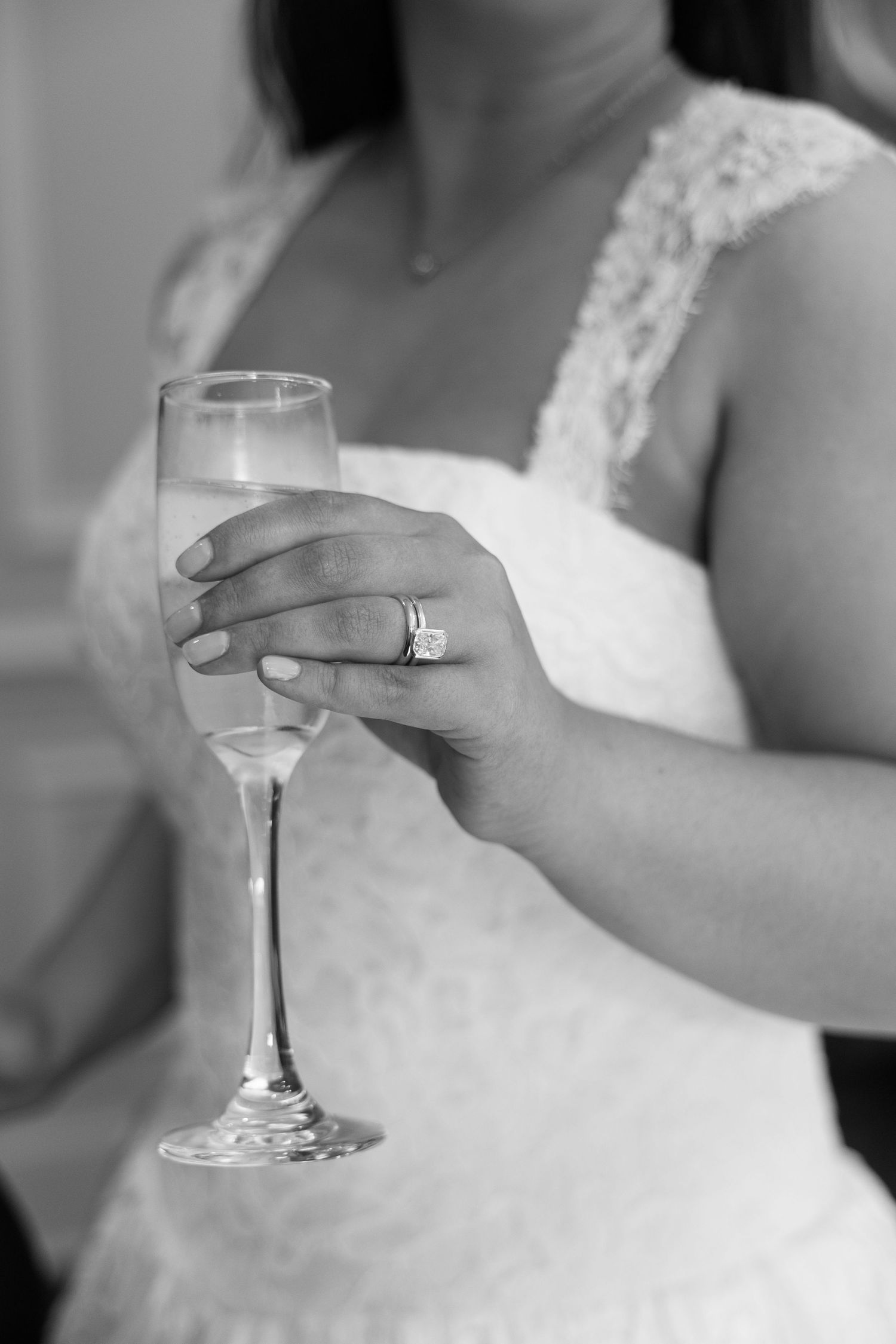 Black and white close-up of a bride holding a champagne flute wearing a diamond wedding ring and lace dress.