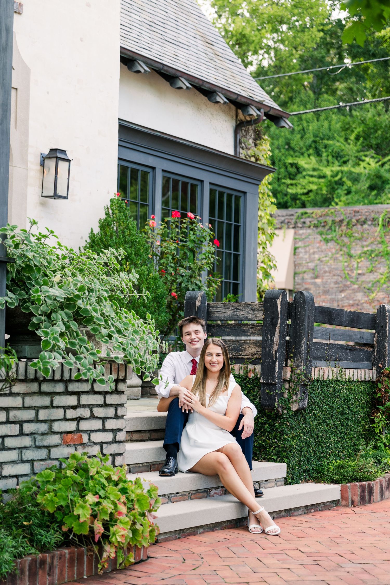 Smiling couple sitting on steps outside charming cottage surrounded by lush greenery and roses.