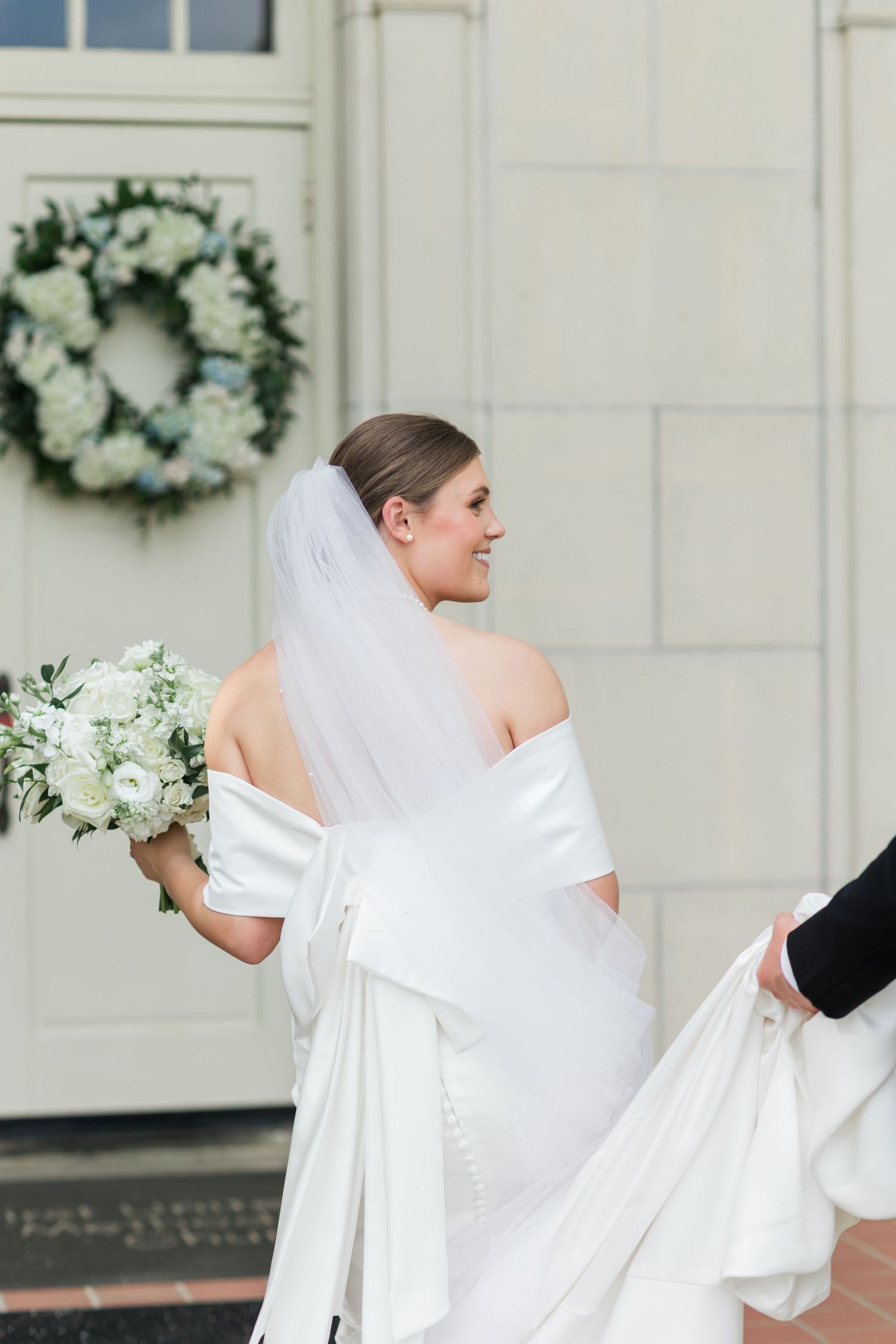 Smiling bride in off-shoulder white gown with bow and veil holds white bouquet outside church with floral wreath.