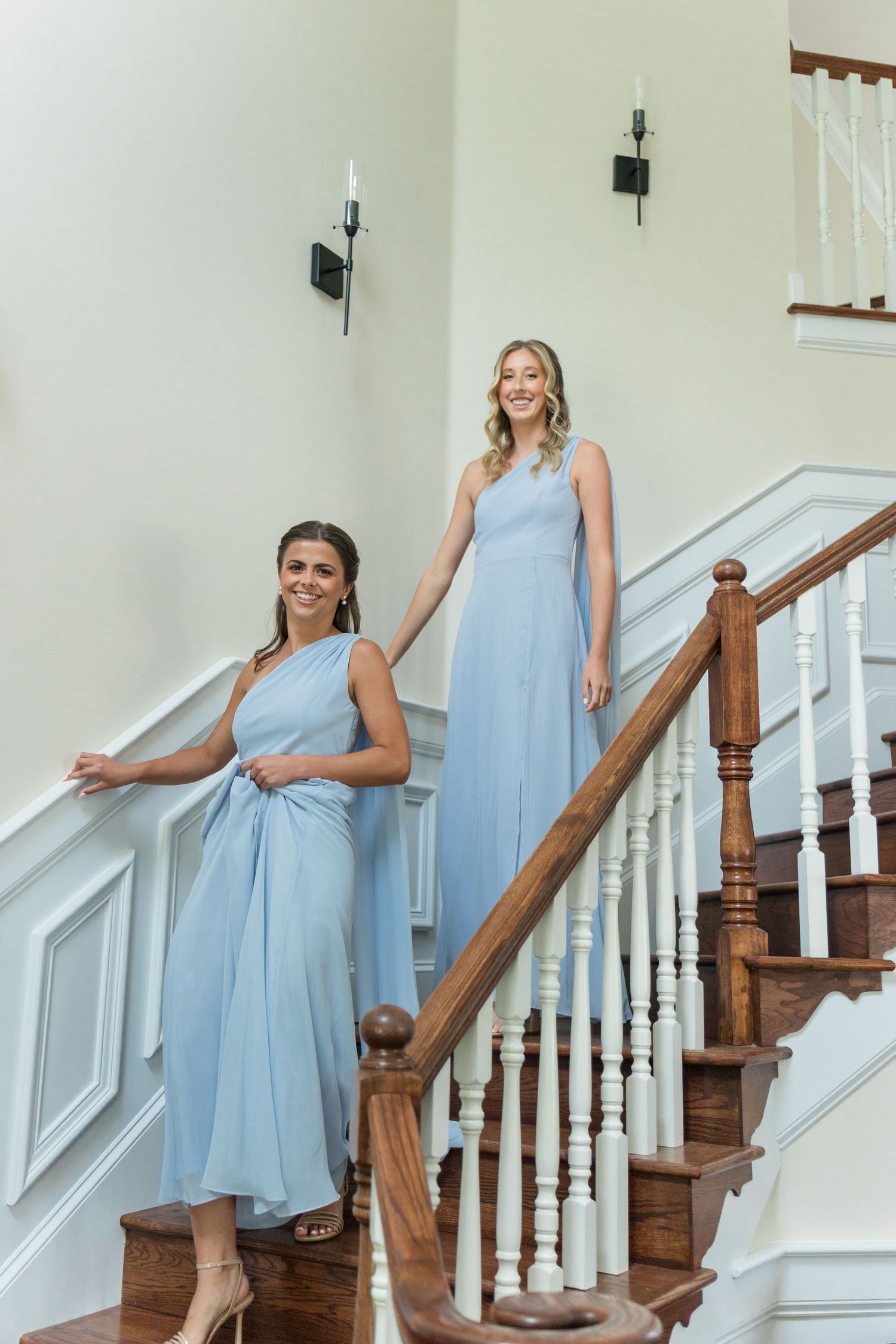 Two bridesmaids in light blue dresses pose elegantly on a grand staircase with white balusters.