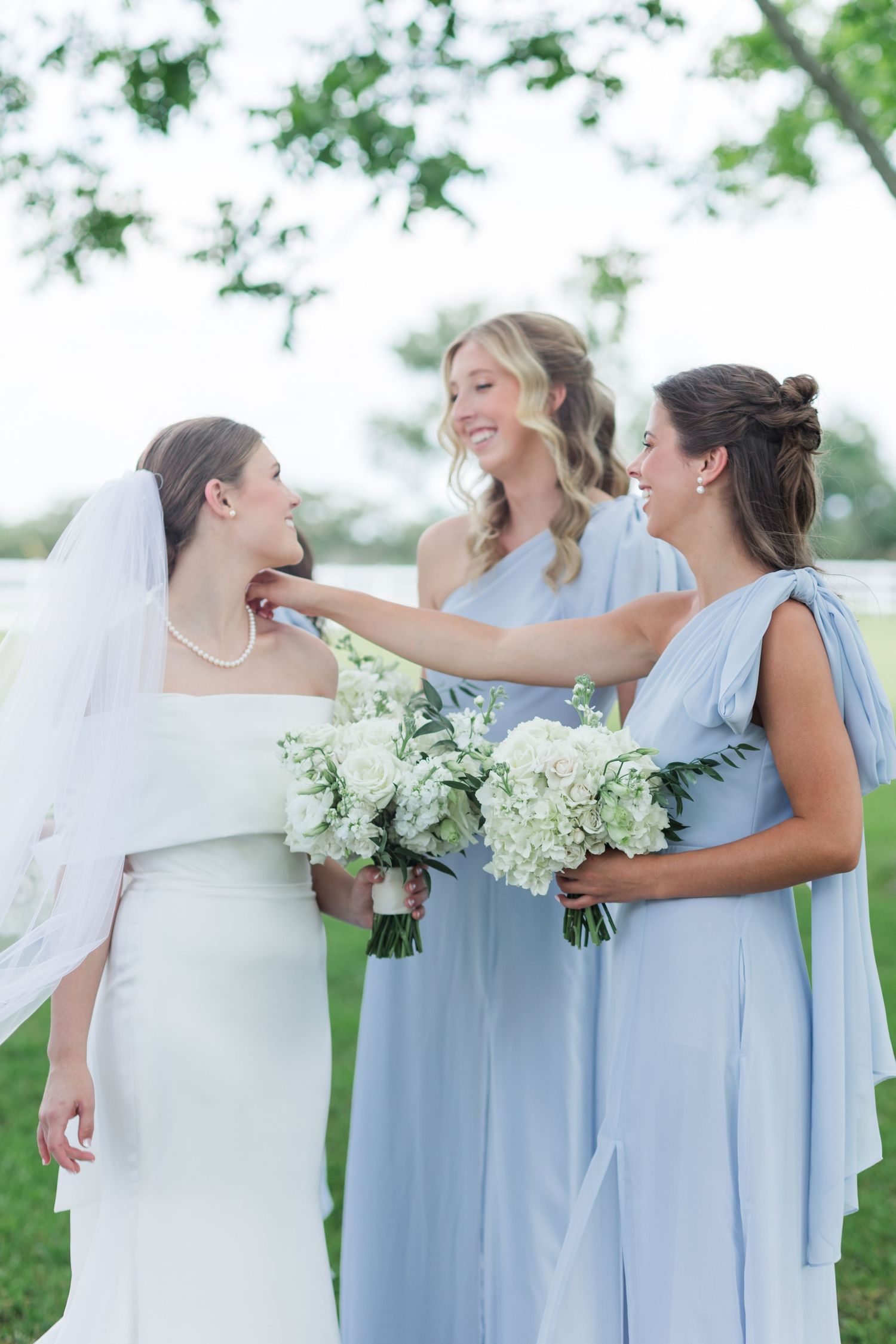 Bride in pearl necklace and veil with two bridesmaids in light blue dresses sharing a joyful moment.