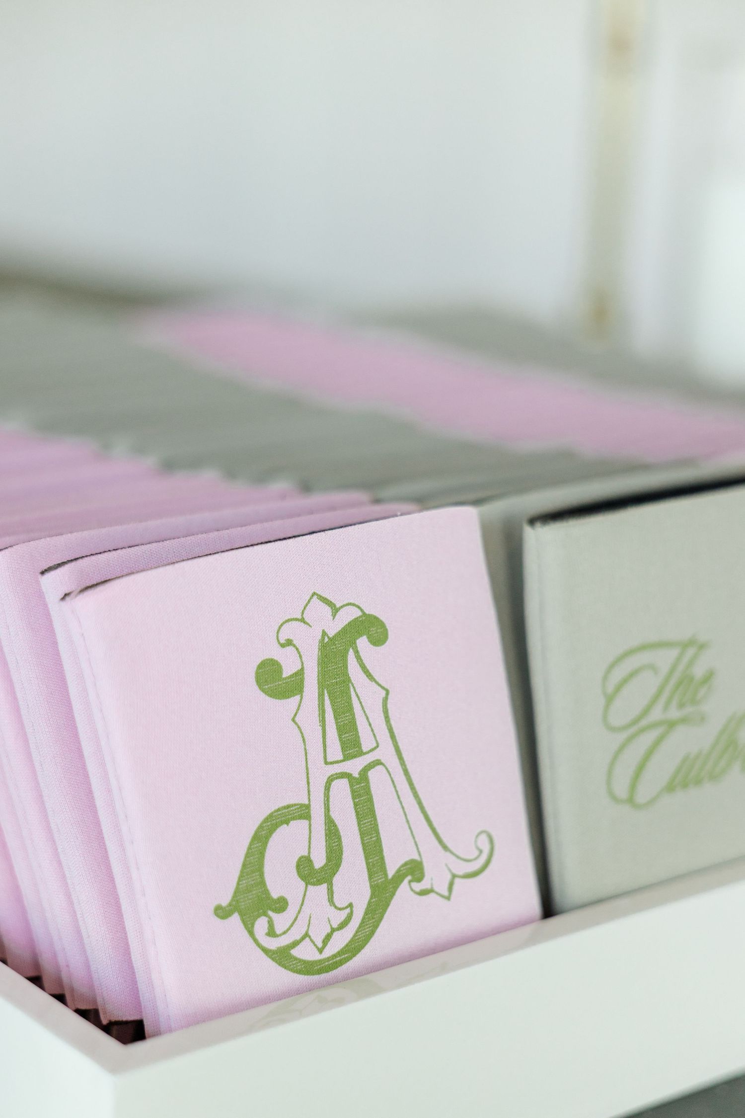 Pink monogrammed napkins with green letter A design displayed in a white box next to gray booklets.