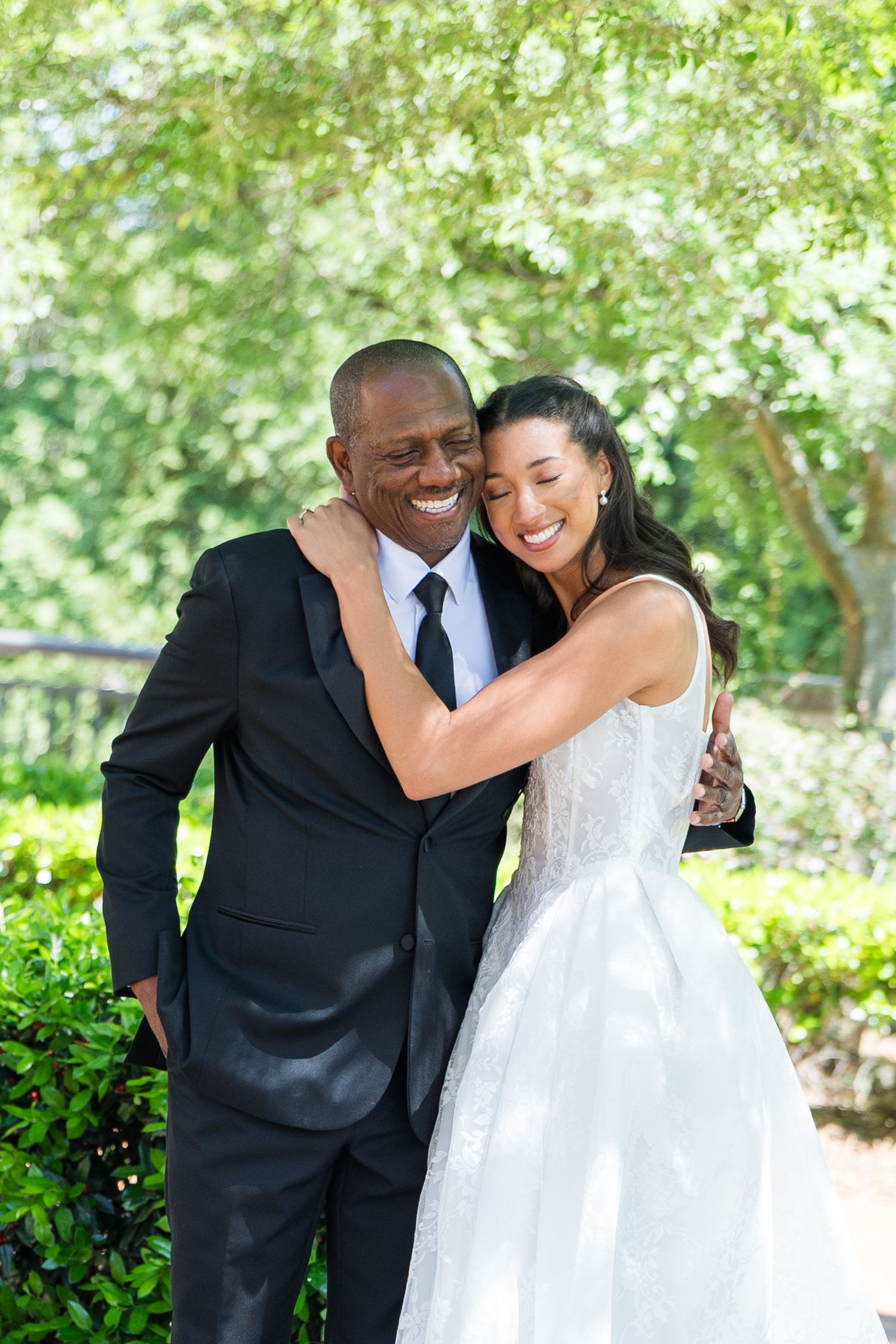 Smiling bride in white gown embraces man in black suit outdoors among lush green trees.