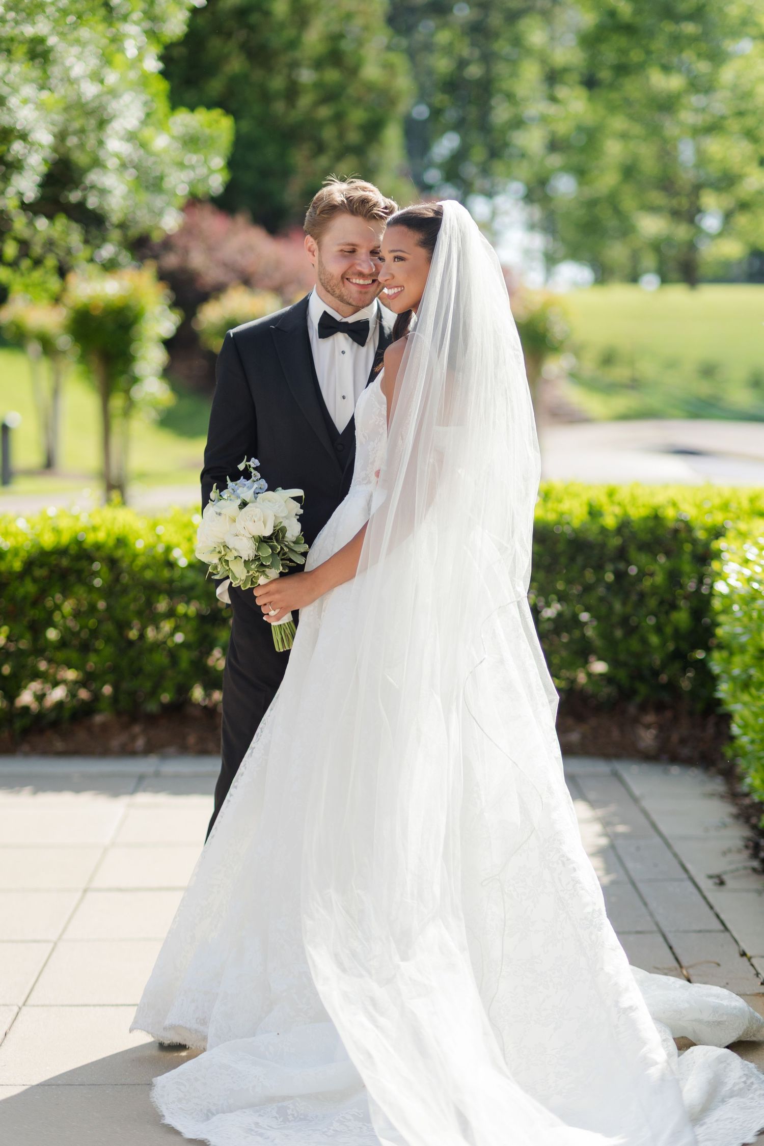 A bride in a white gown and veil holds a bouquet while embracing a groom in a black tuxedo outdoors.
