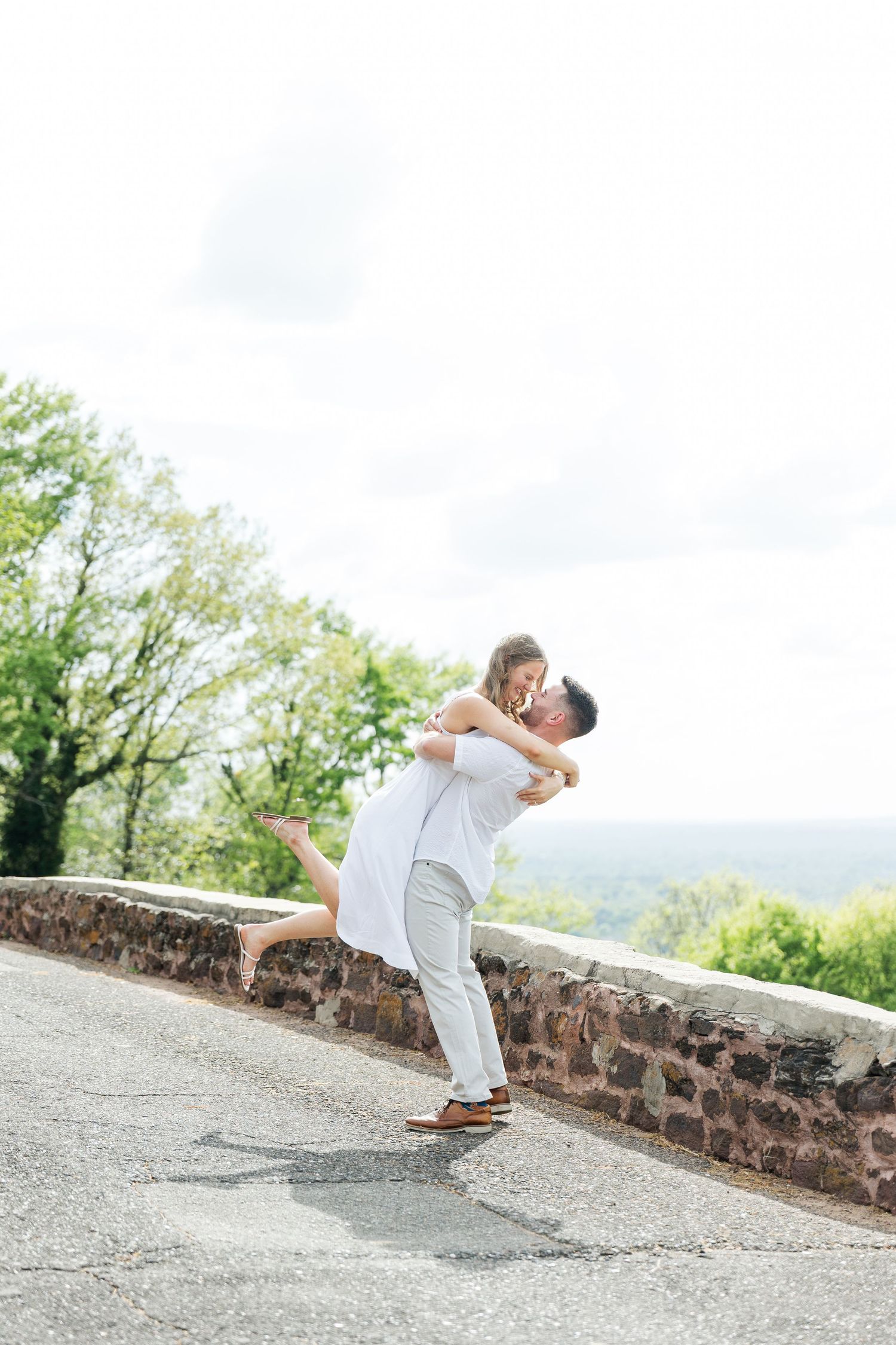 Romantic couple embracing outdoors, man lifting woman in white dress near stone wall with green trees background.