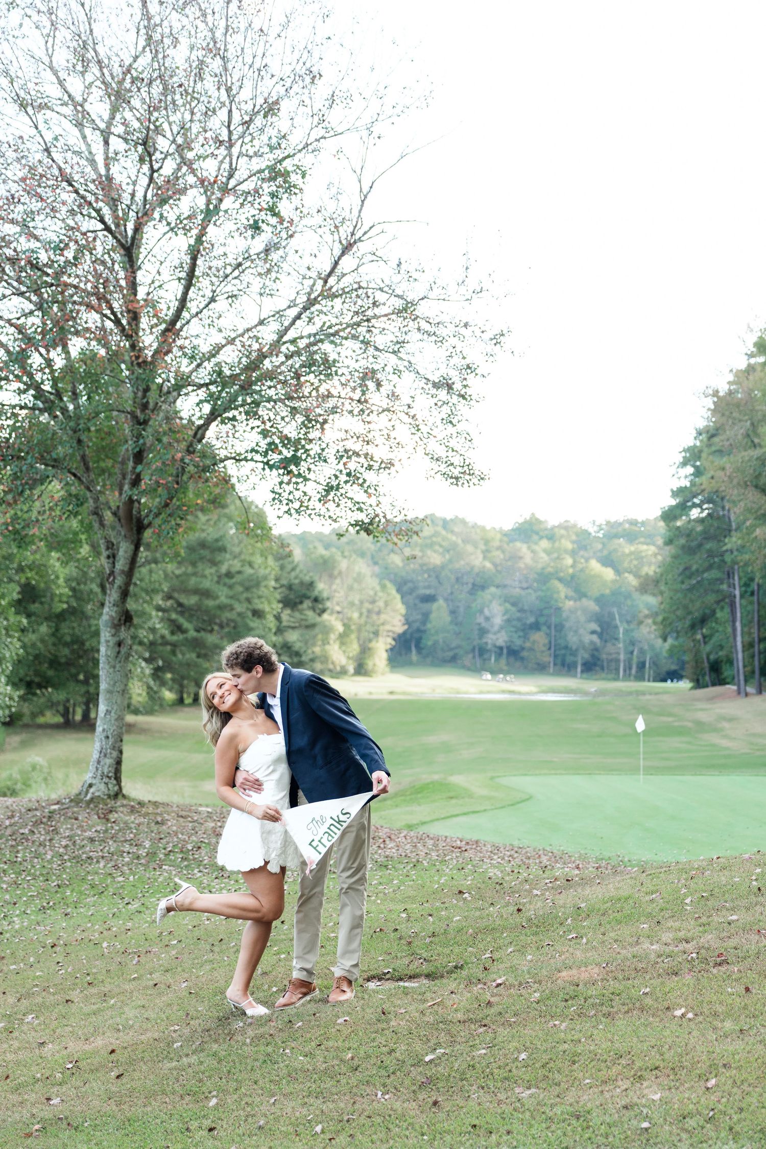 Couple kissing and laughing on a golf course, bride in white mini dress holding an engagement pennant sign.