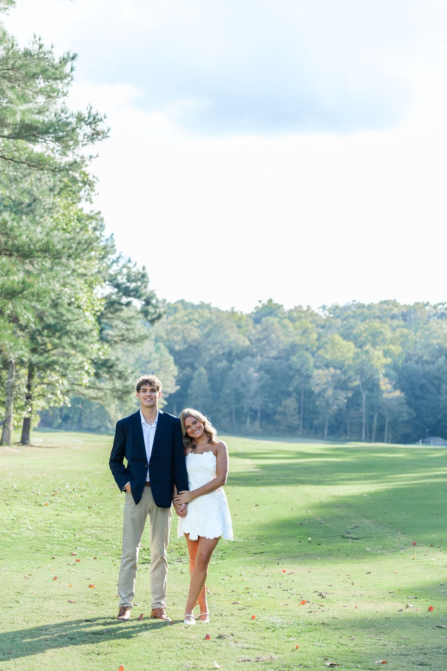 A couple poses on a lush green golf course, man in navy blazer and woman in white dress, surrounded by tall trees.