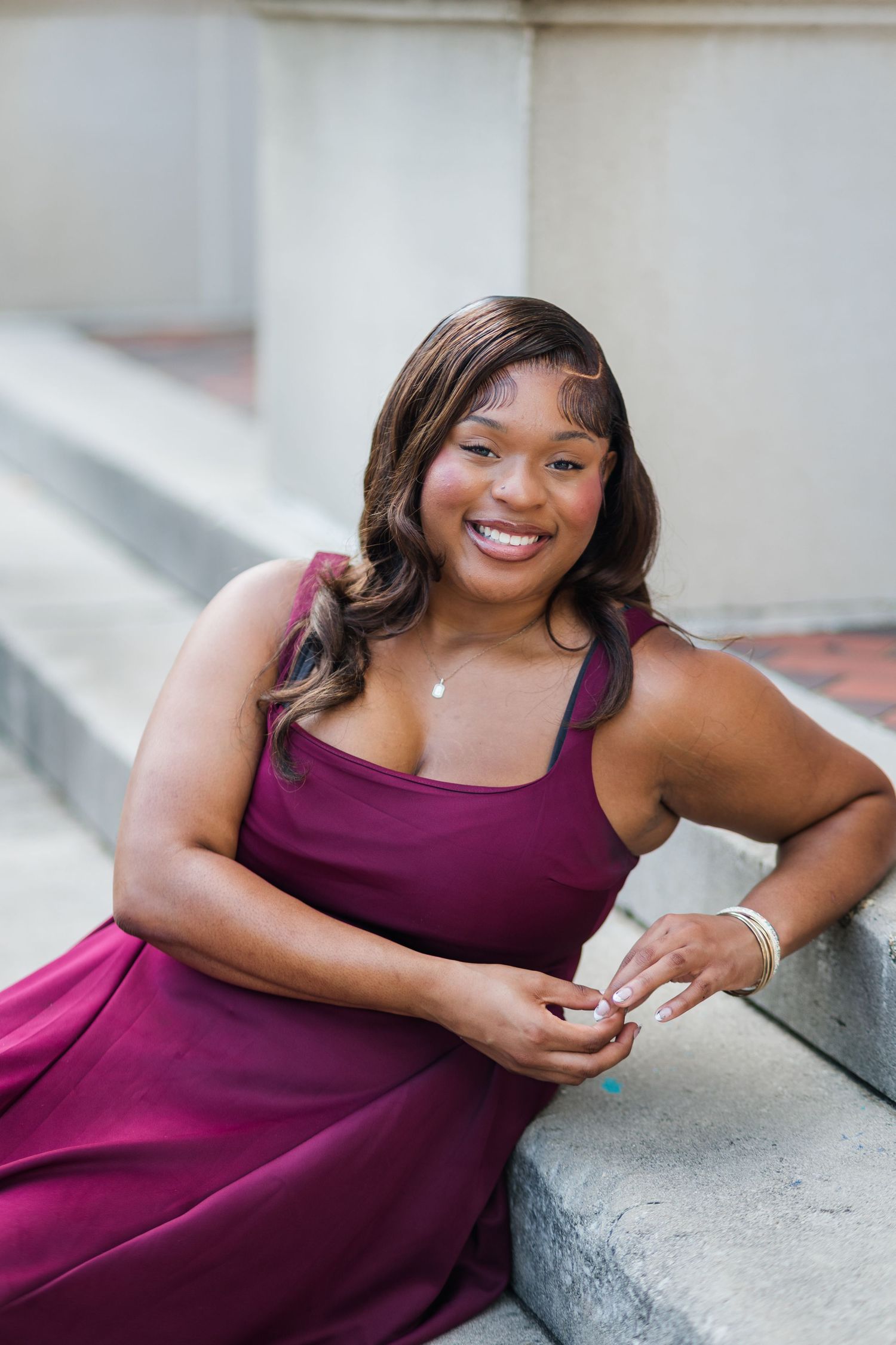 Smiling young woman in burgundy dress sitting on stone steps during graduation photo session.