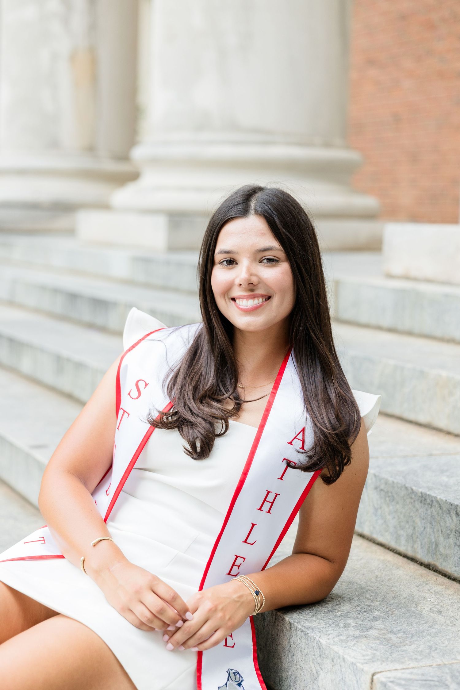 Smiling female graduate in white dress with Student Athlete sash sits on steps of brick university building.