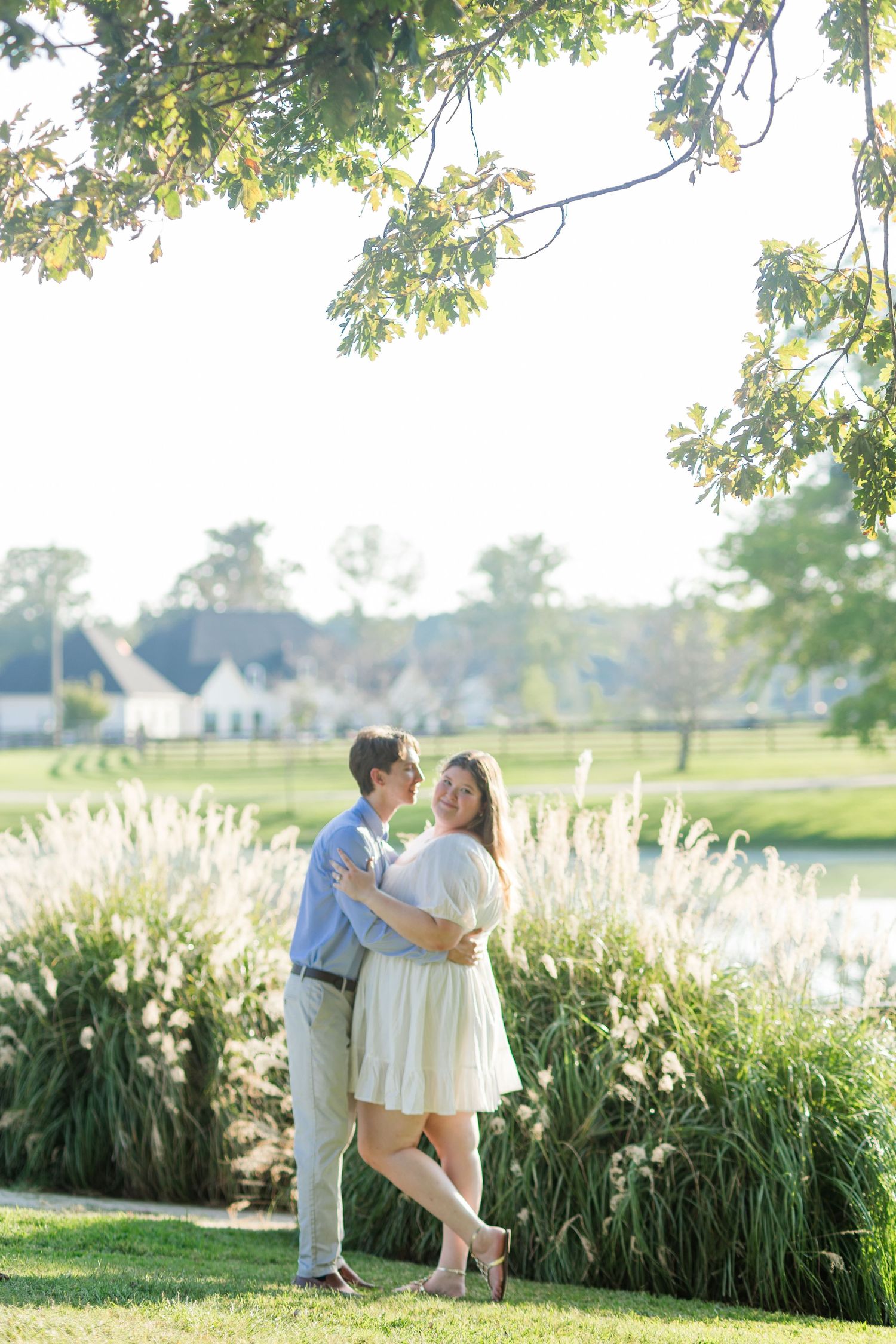 Couple embracing and smiling at each other in a sunlit park with ornamental grasses and a pond in the background.