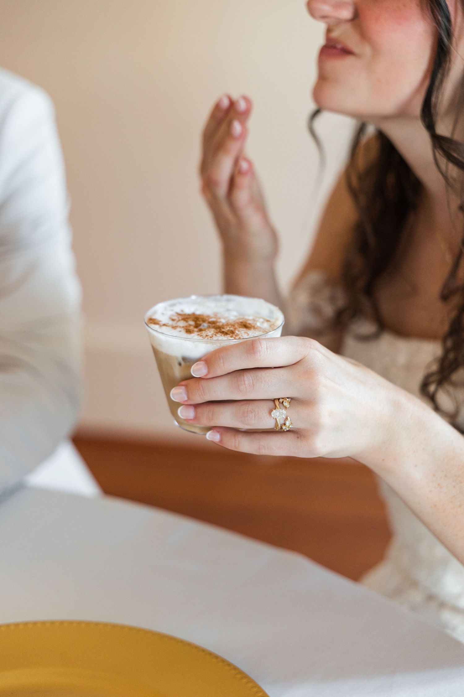 Close-up of a bride holding a frothy cappuccino coffee drink while wearing a gold engagement ring at a cafe.