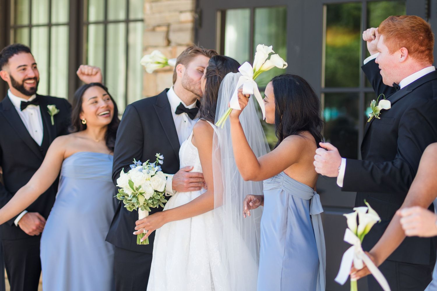 Wedding party celebrating as bride and groom kiss, bridesmaids in blue dresses holding white bouquets, groomsmen in black tuxedos.