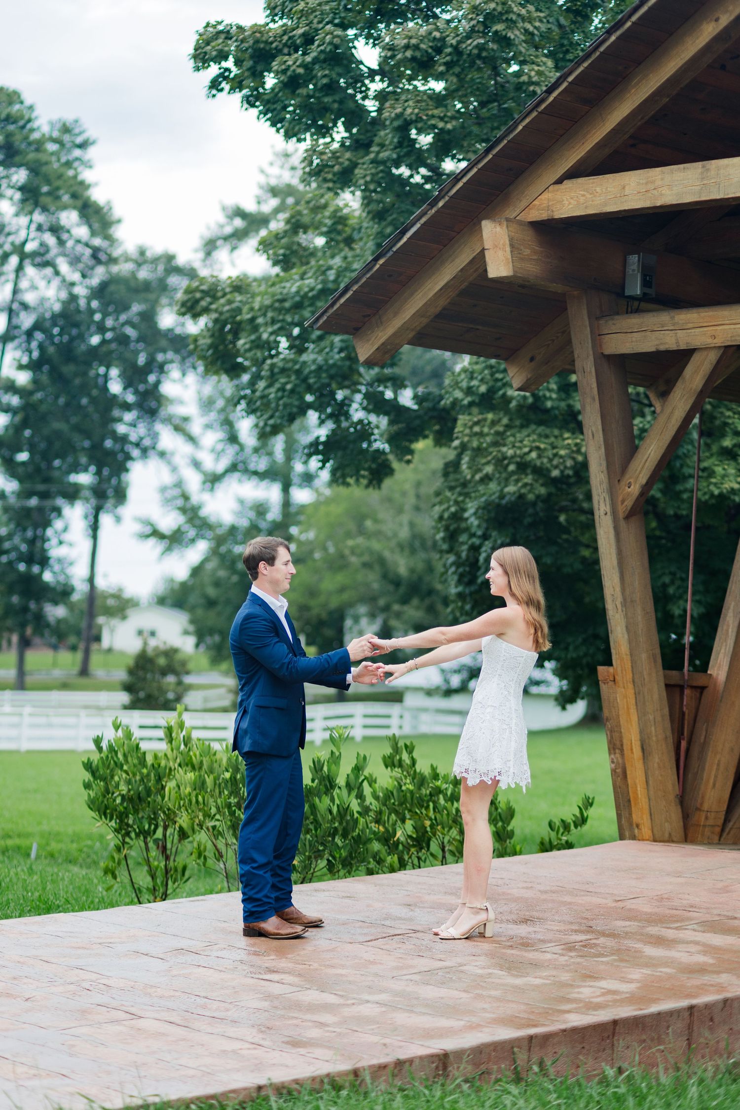 Couple holding hands on outdoor wooden pavilion dance floor, man in navy suit, woman in white dress, green trees behind.