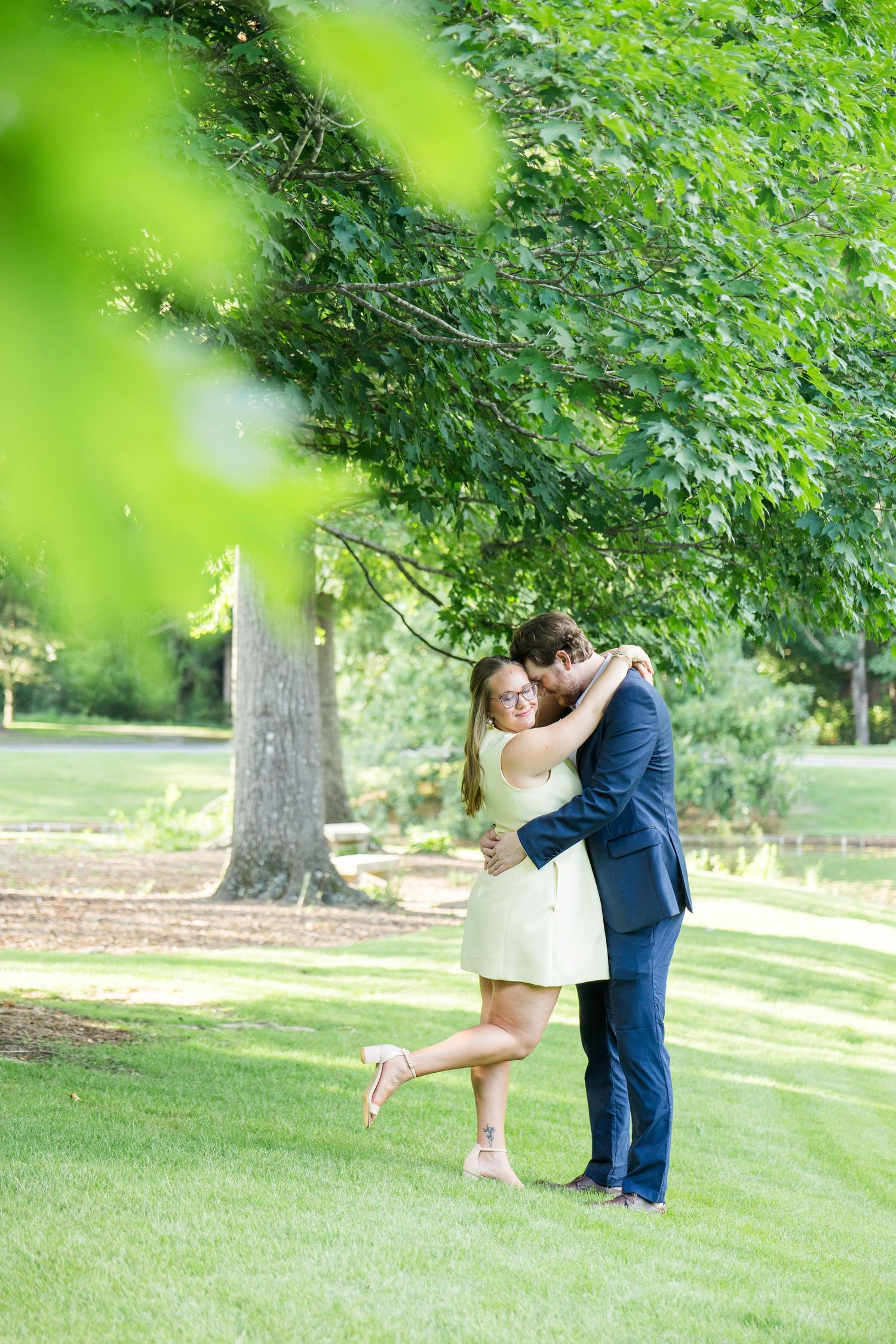 Couple embracing and laughing in a lush green park, woman in yellow dress lifting her foot playfully.