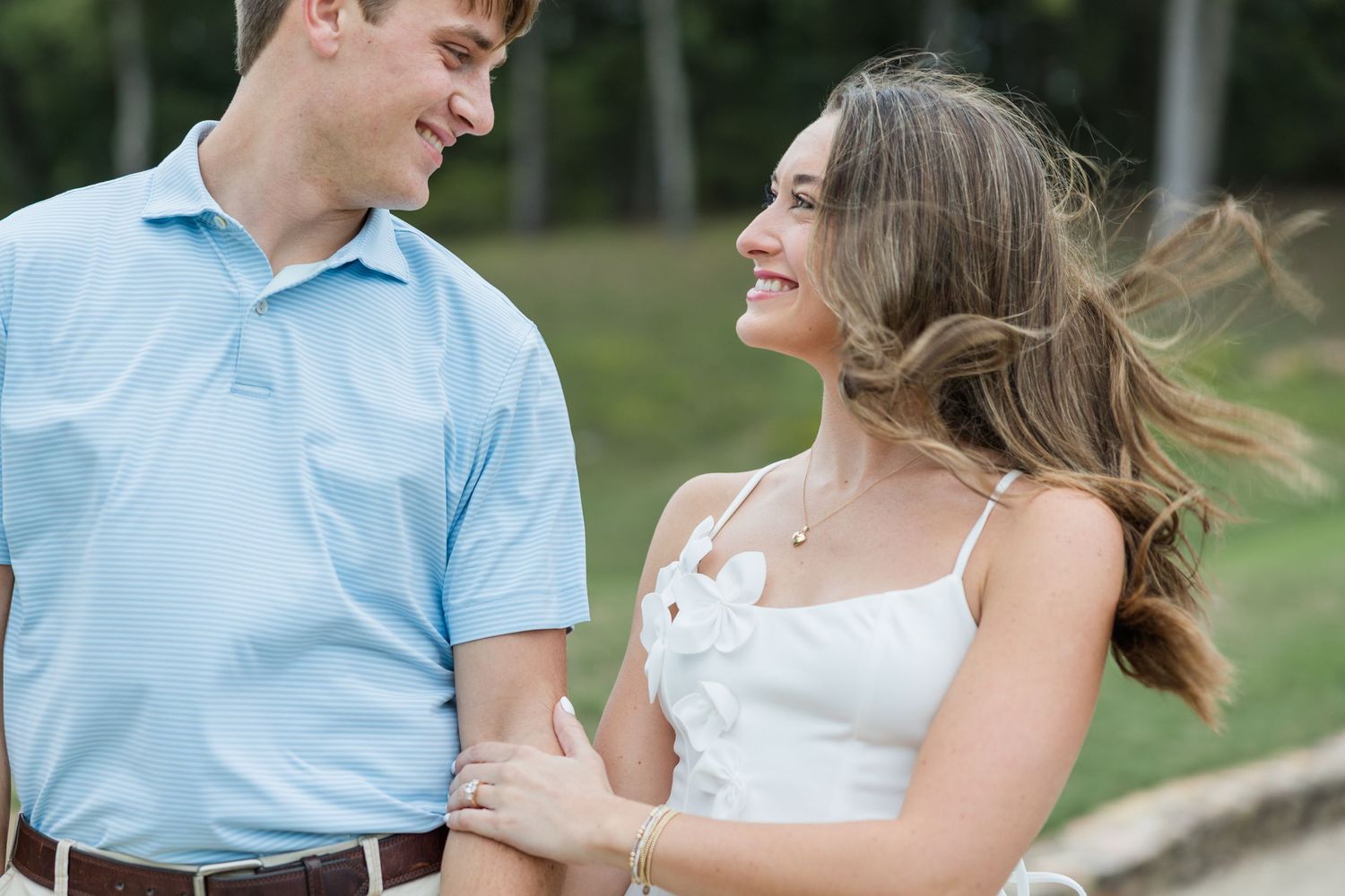 Smiling couple standing close outdoors, a man in a light blue polo and a woman in a white top with windblown hair, in a green park setting.