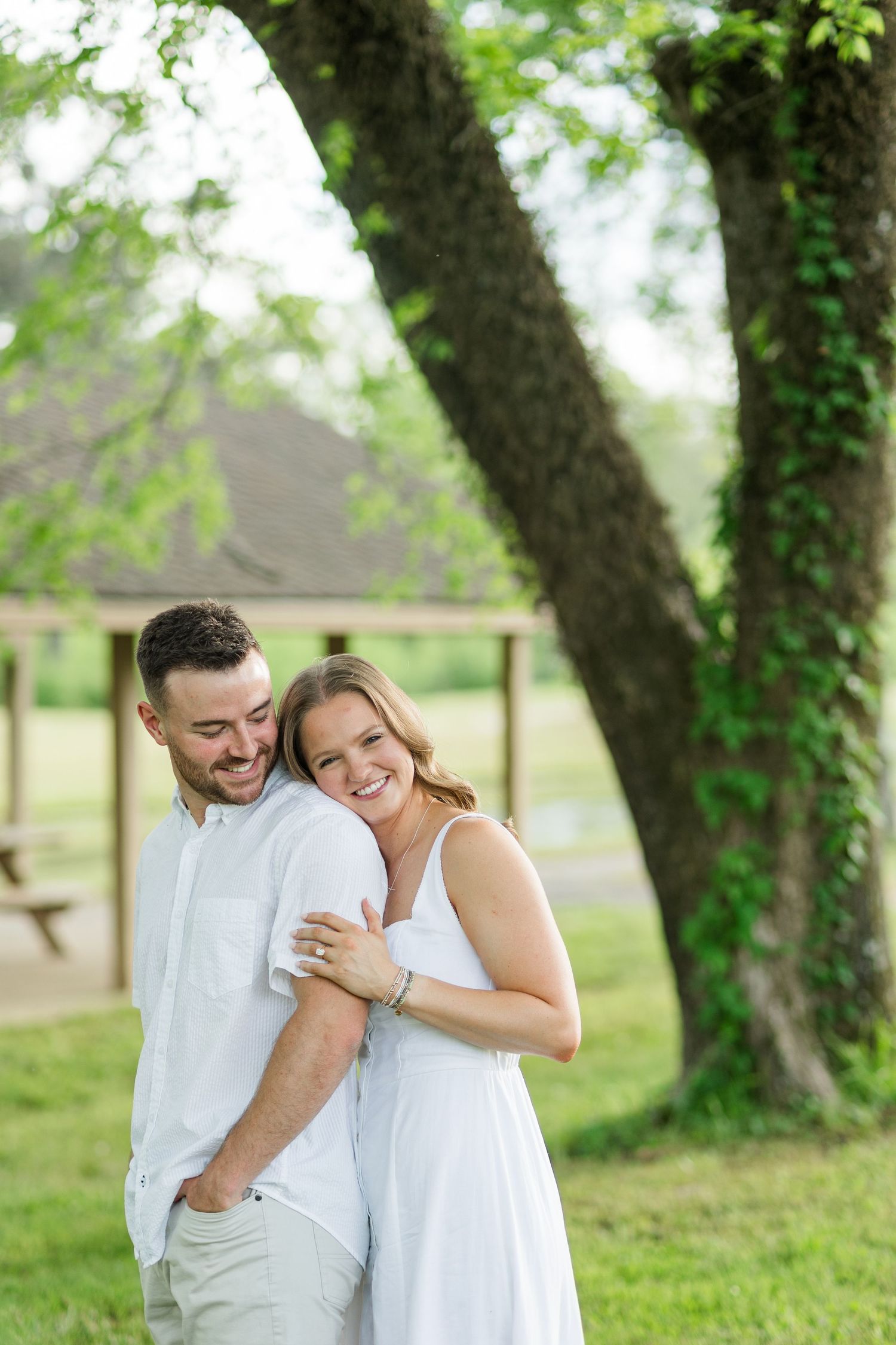 Happy couple embracing outdoors under a large tree, both wearing white outfits in a lush green park setting.