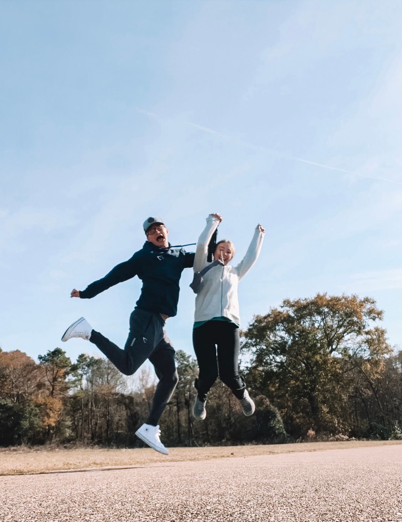 Joyful couple jumping in air against blue sky with trees in outdoor park setting.