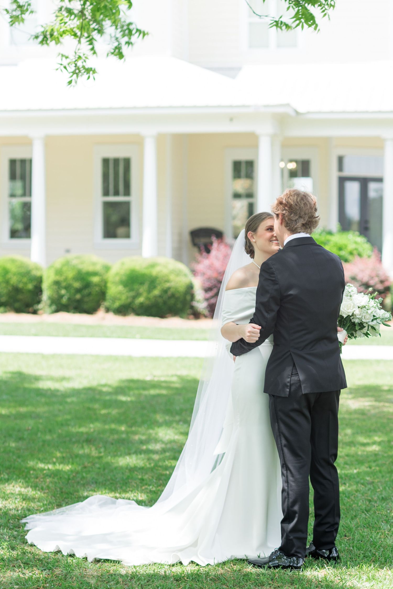 Bride and groom share romantic first look moment on lush green lawn before white colonial estate.