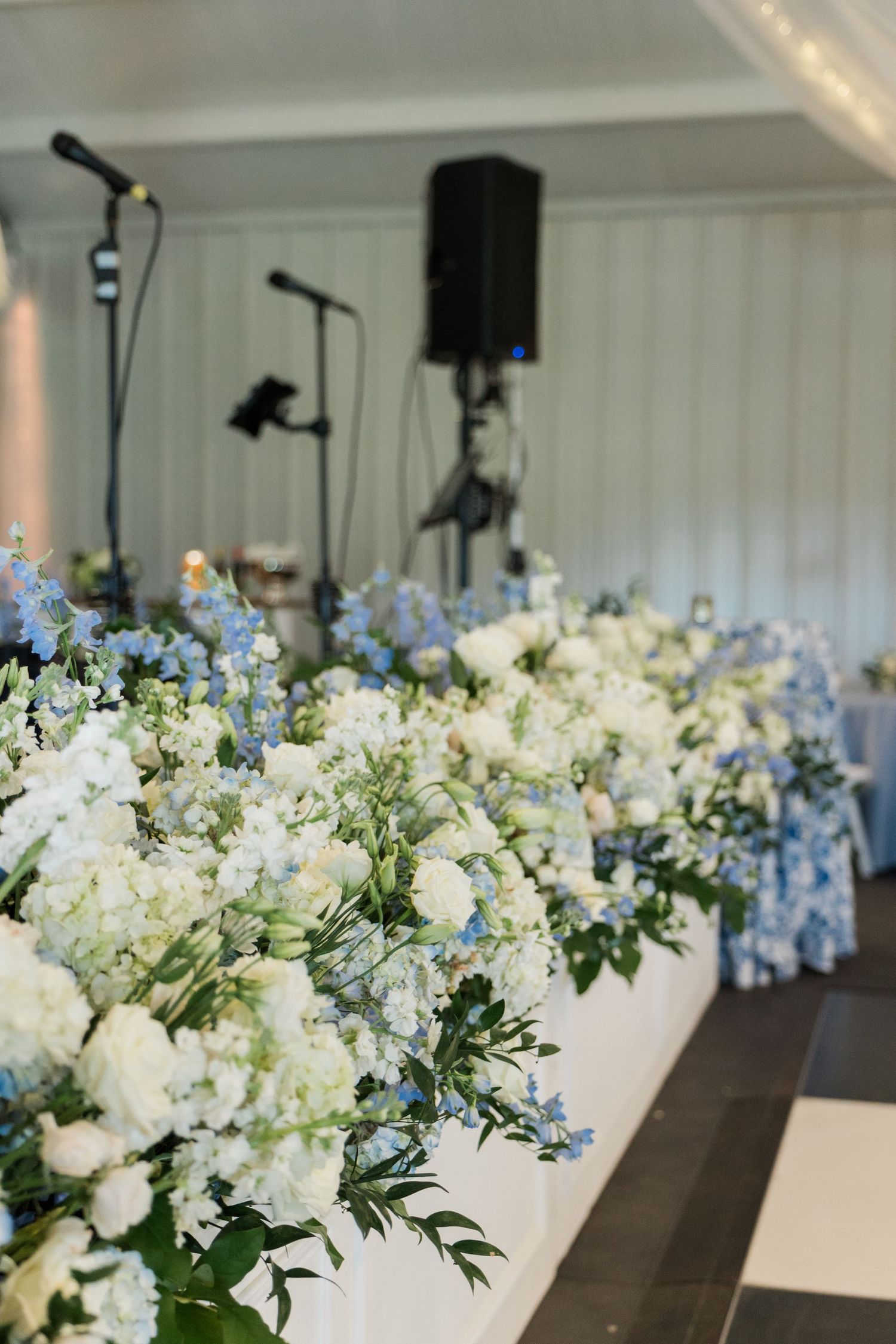 Elegant wedding reception table adorned with lush white and blue floral arrangements with greenery in a white tent.