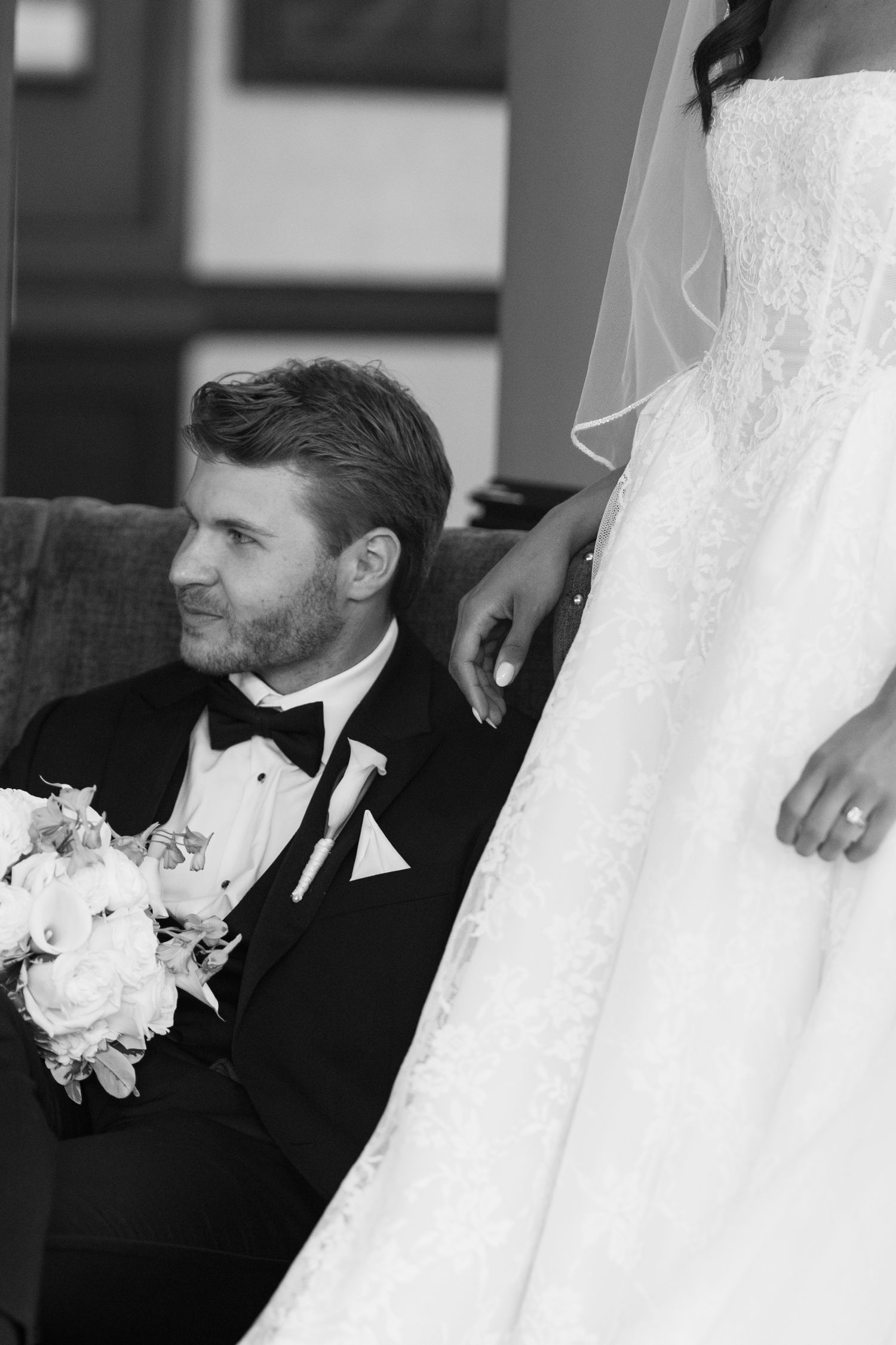 Black and white wedding photo of groom in tuxedo holding bouquet seated beside bride in lace gown with veil.