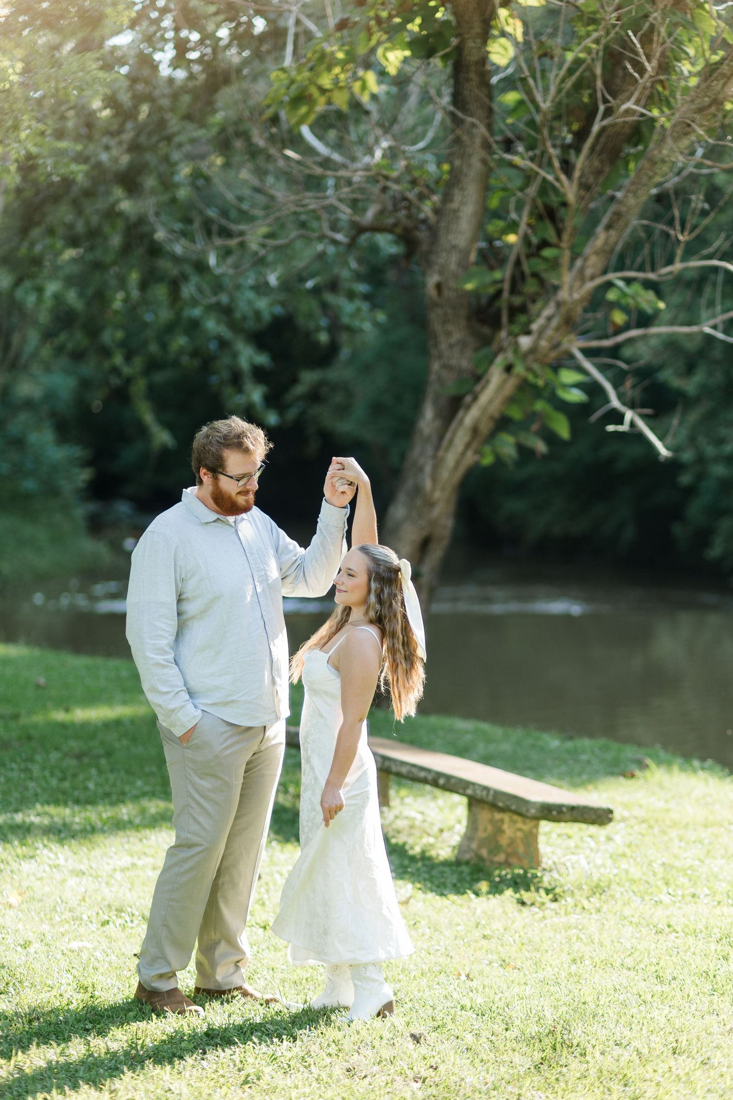 Couple dancing outdoors near river, man spinning woman in white dress on green grass under trees.