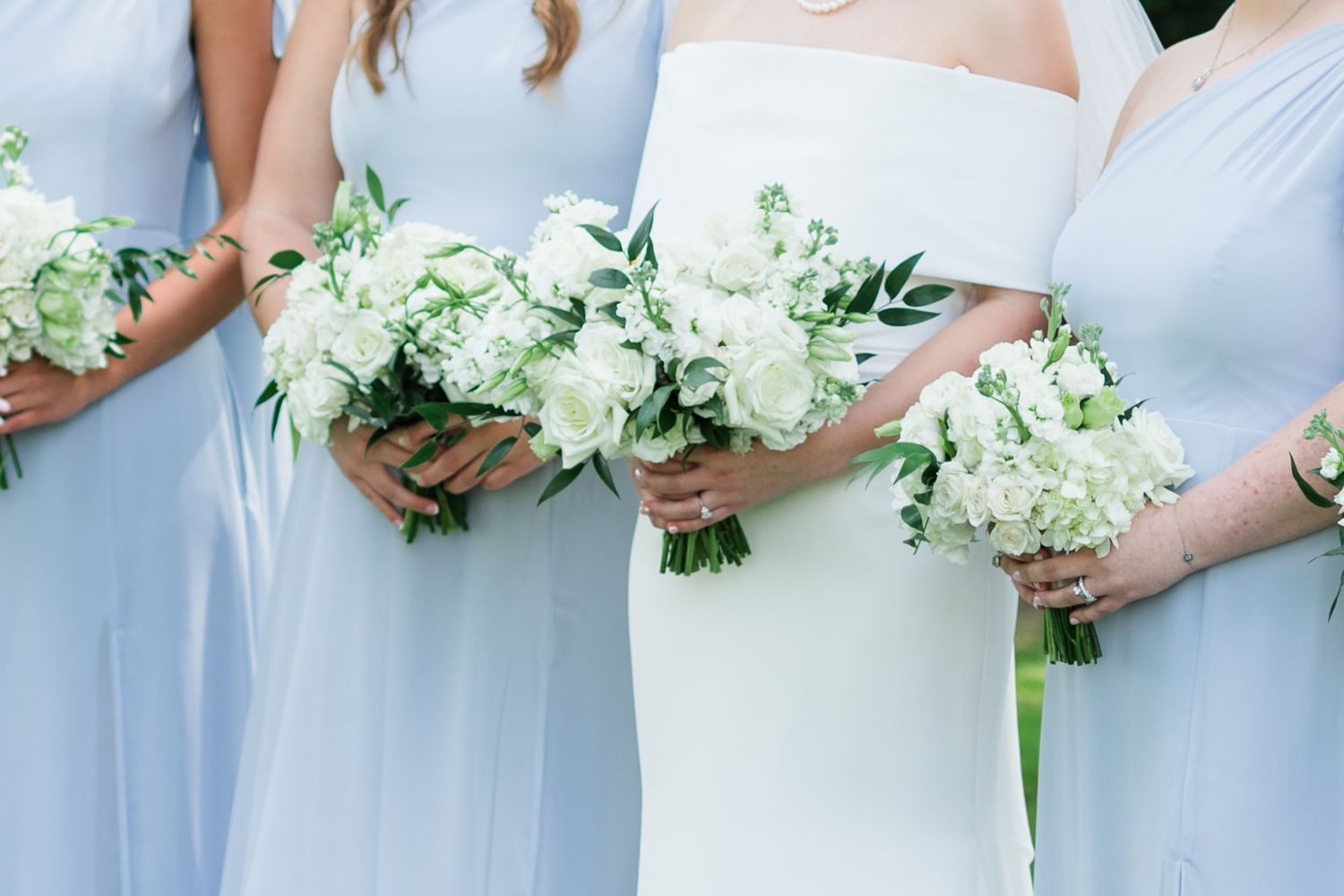 Bride in white gown and bridesmaids in light blue dresses holding white floral bouquets at a wedding ceremony.