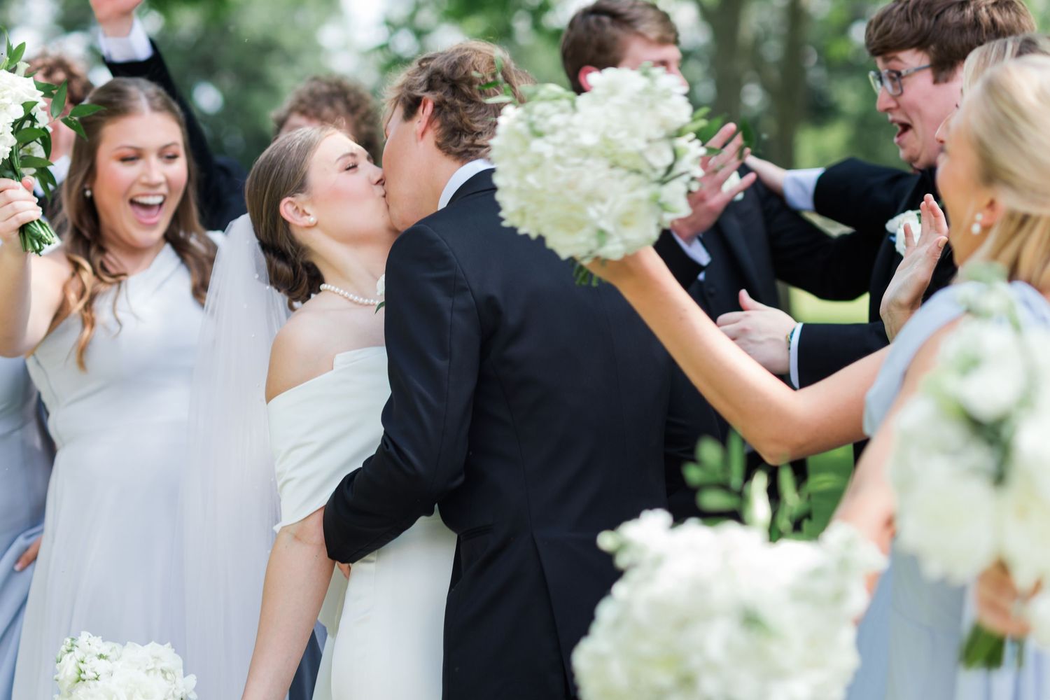 Bride and groom sharing a kiss surrounded by cheering wedding party holding white bouquets.