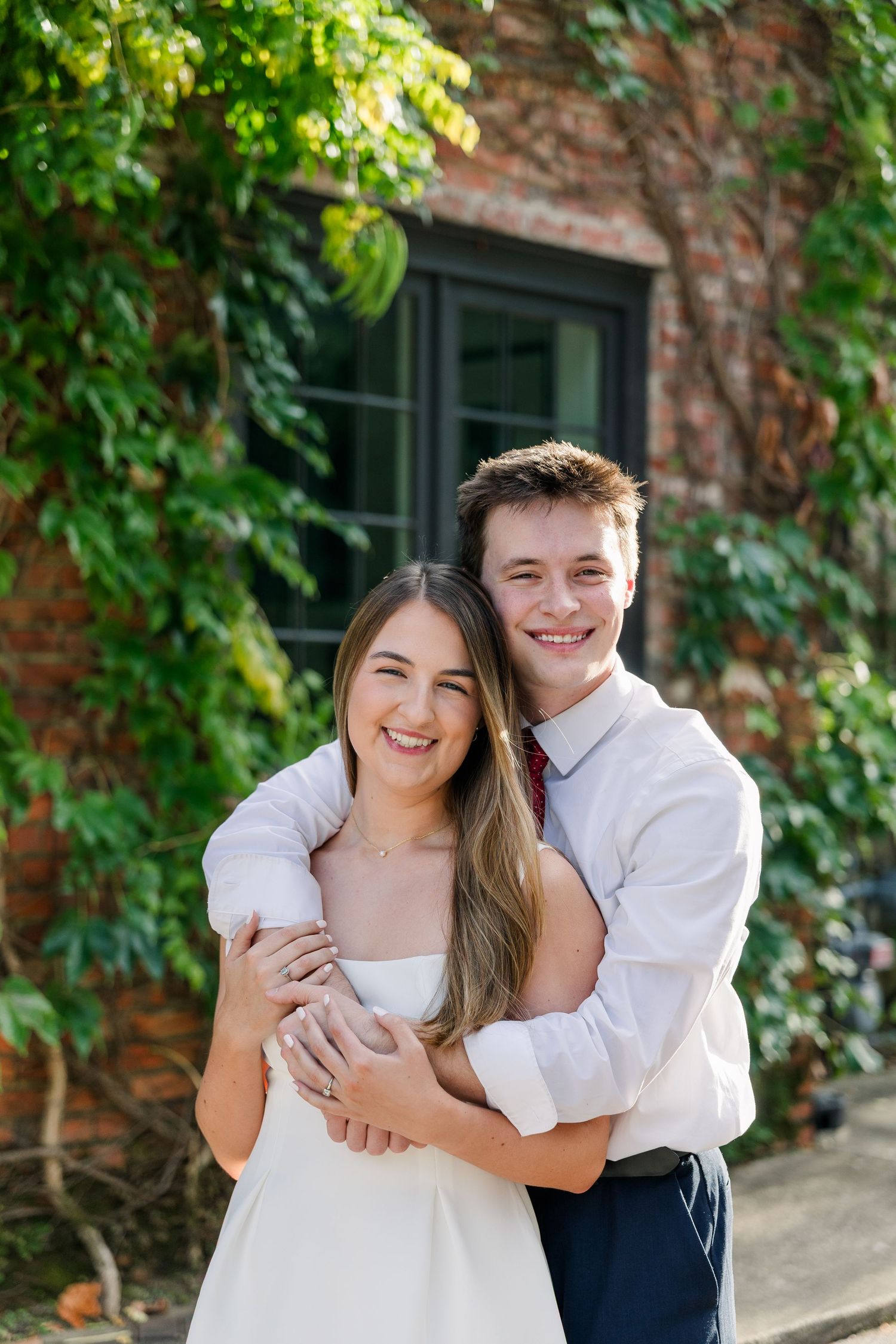 A smiling couple embraces outdoors in front of a brick wall covered in lush green ivy near a dark-framed window.