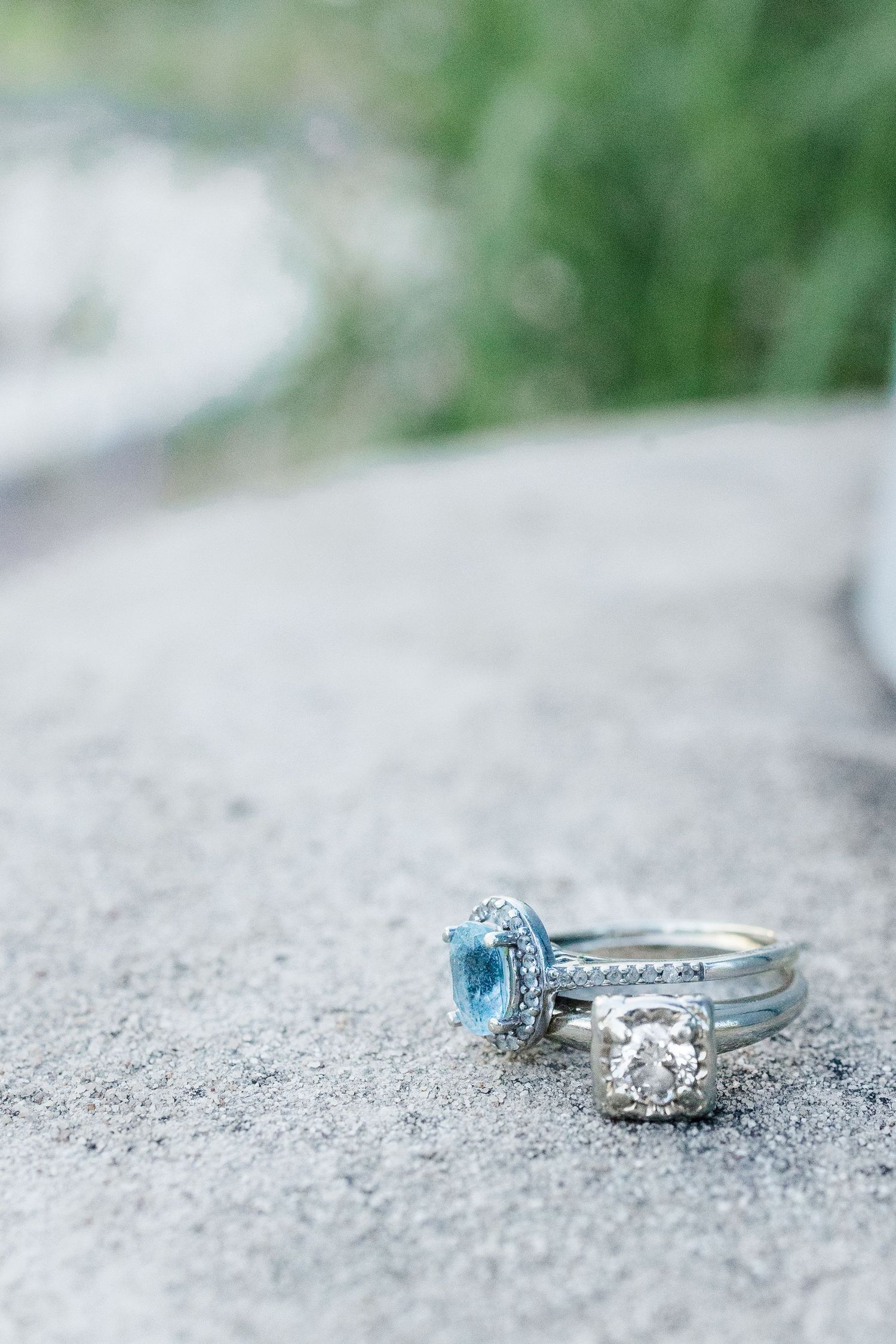 Two elegant silver rings with blue topaz and diamond halo settings displayed on a stone surface outdoors.