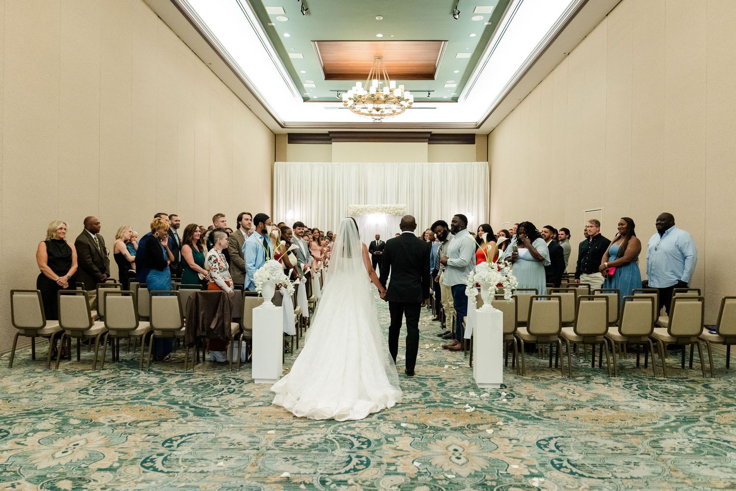Bride and groom stand at the altar during an indoor wedding ceremony in an elegant ballroom with seated guests.