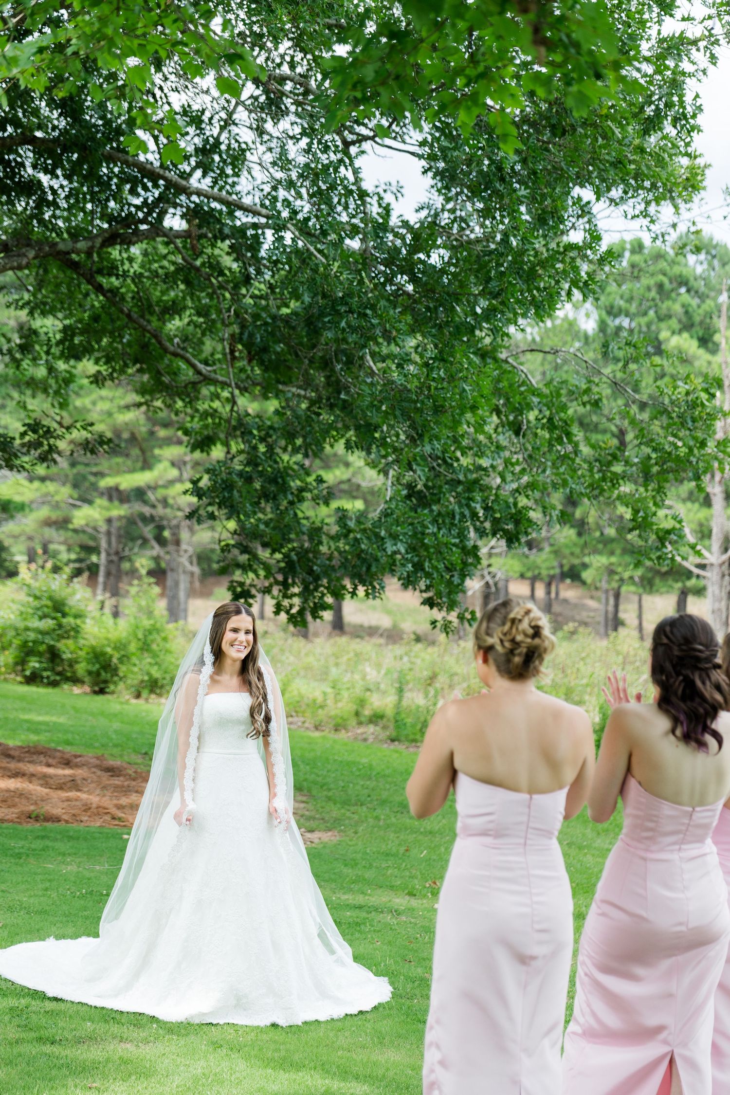 Bride in white gown and veil smiles at bridesmaids in pink dresses on a lush green lawn.