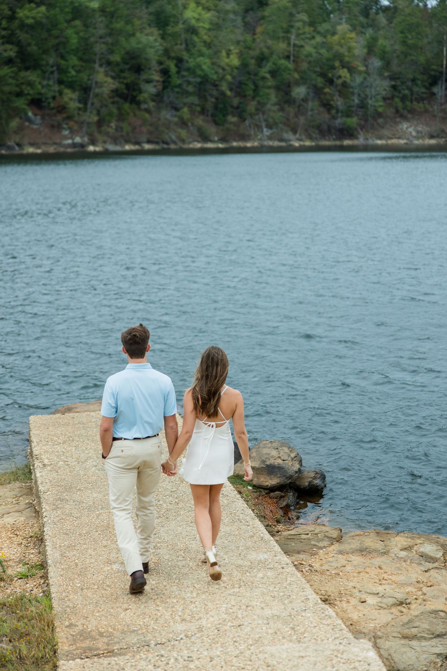 A couple walks hand in hand along a stone path toward a calm lake surrounded by a dense green forest.