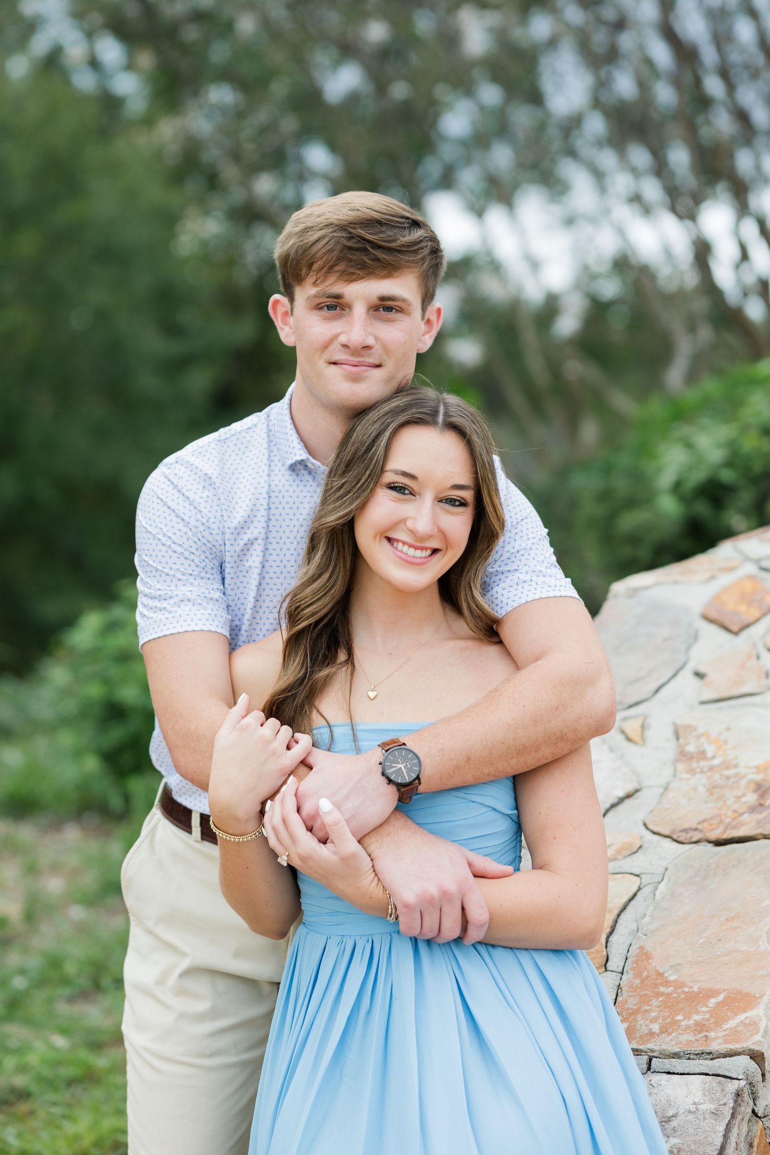 Smiling couple in an embrace outdoors, the young man hugging a woman in a blue dress from behind beside a stone wall.