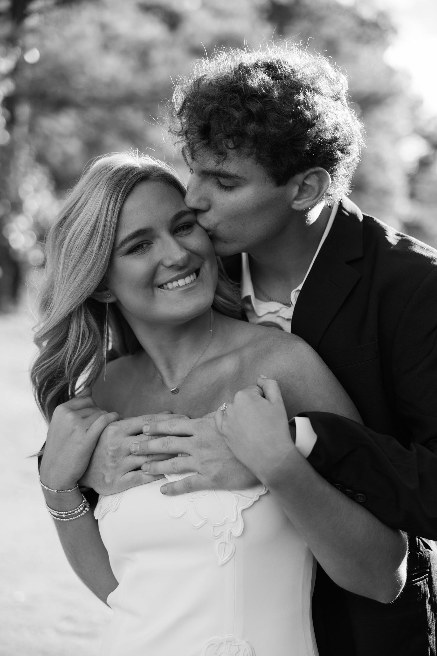 Groom kissing bride on the cheek as she smiles in a white dress during an outdoor engagement session.