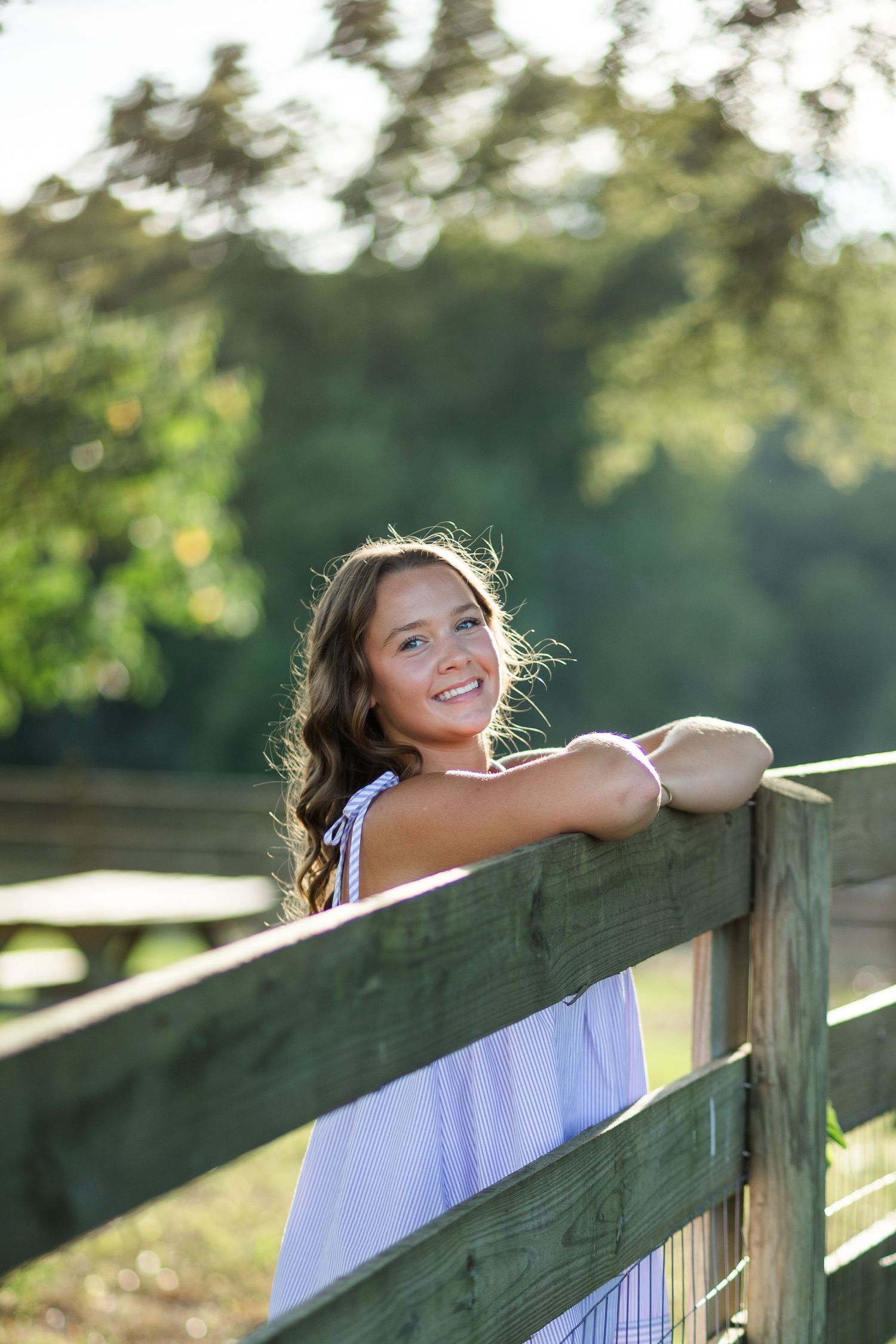 A smiling young girl with long brown hair leans on a wooden fence outdoors, wearing a white dress on a sunny day.