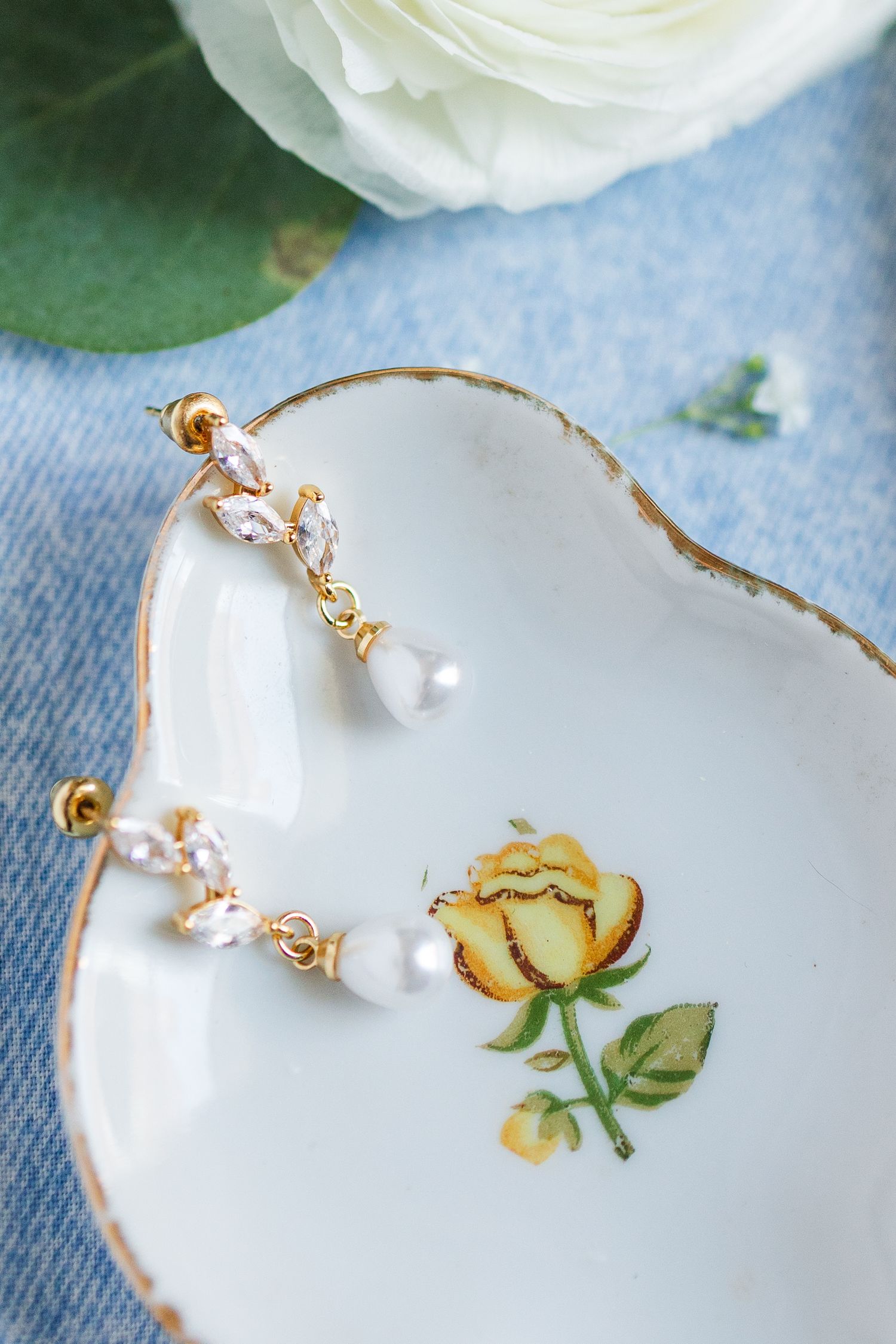 Gold drop earrings with crystal and pearl details displayed on vintage floral porcelain dish with white rose.