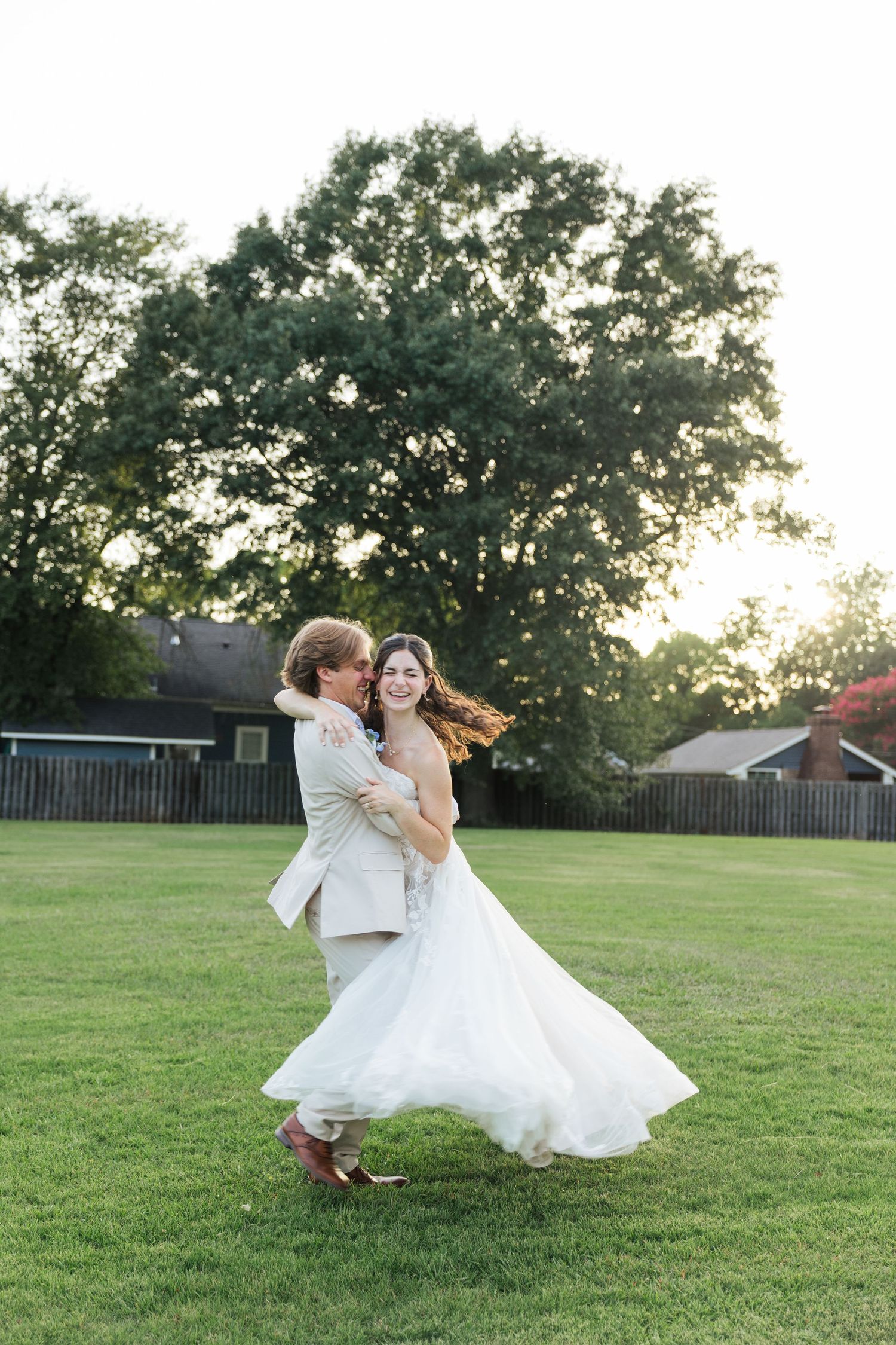 Bride and groom dancing joyfully on green lawn at golden hour, groom lifting bride in white flowing dress.