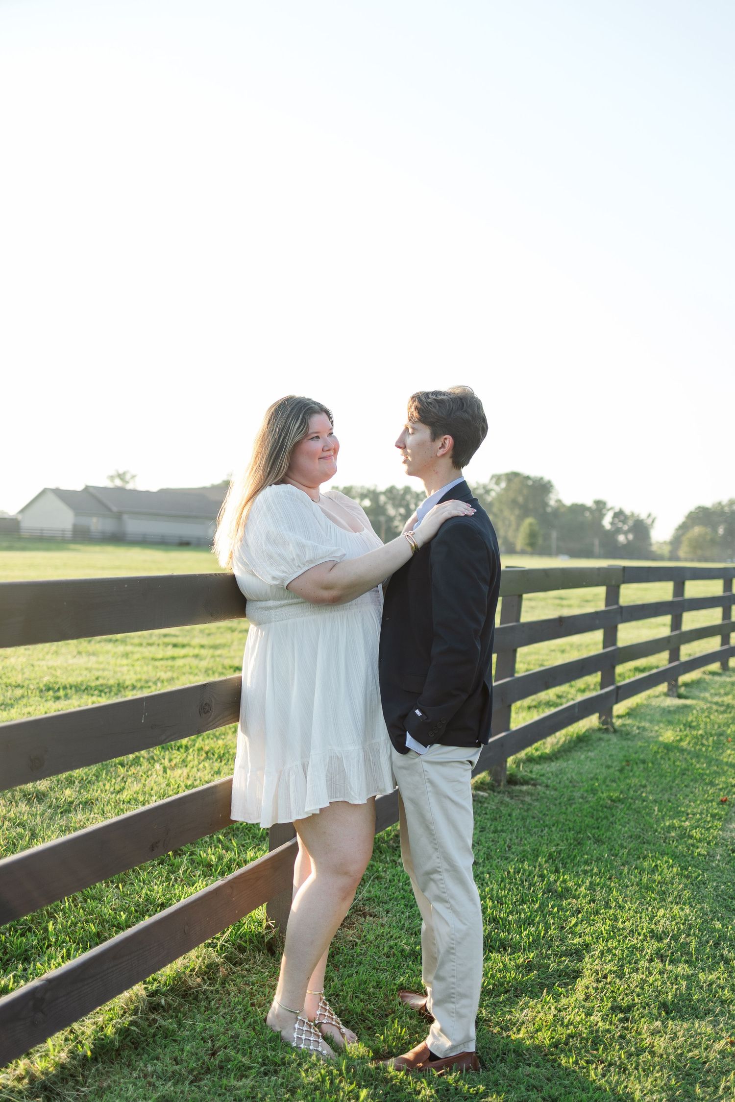 Couple gazing at each other near a wooden fence on a sunny farm during engagement session.