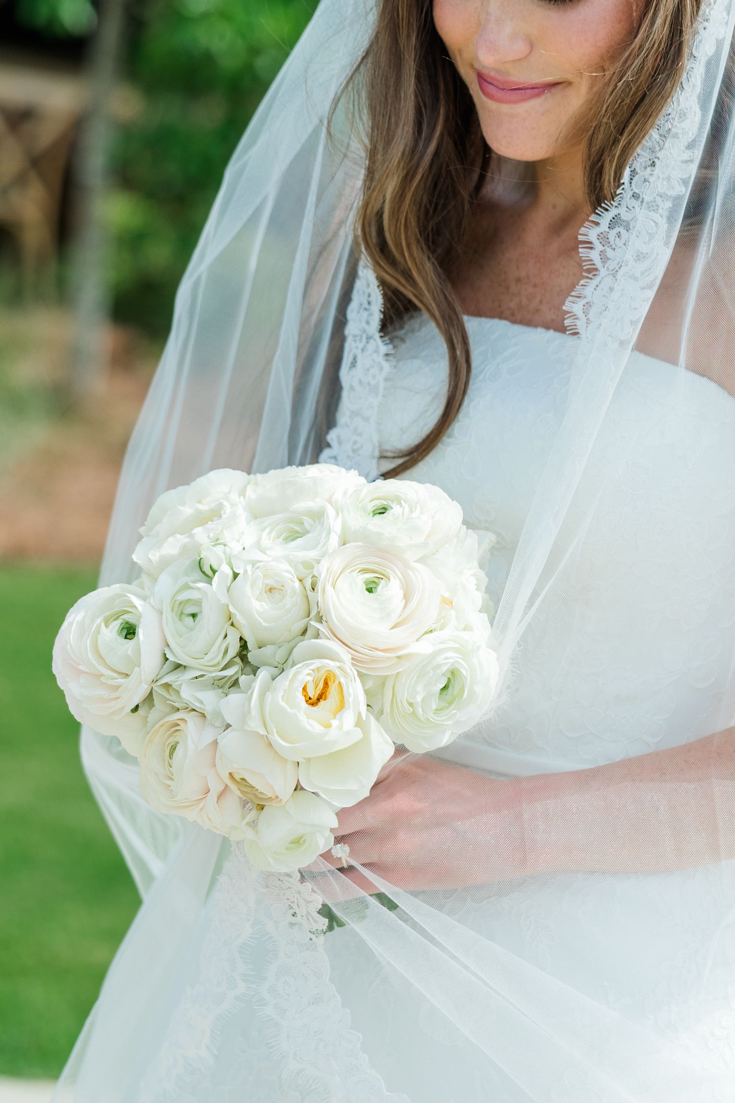 Bride in white lace wedding dress and veil holding a round bouquet of white ranunculus flowers outdoors.