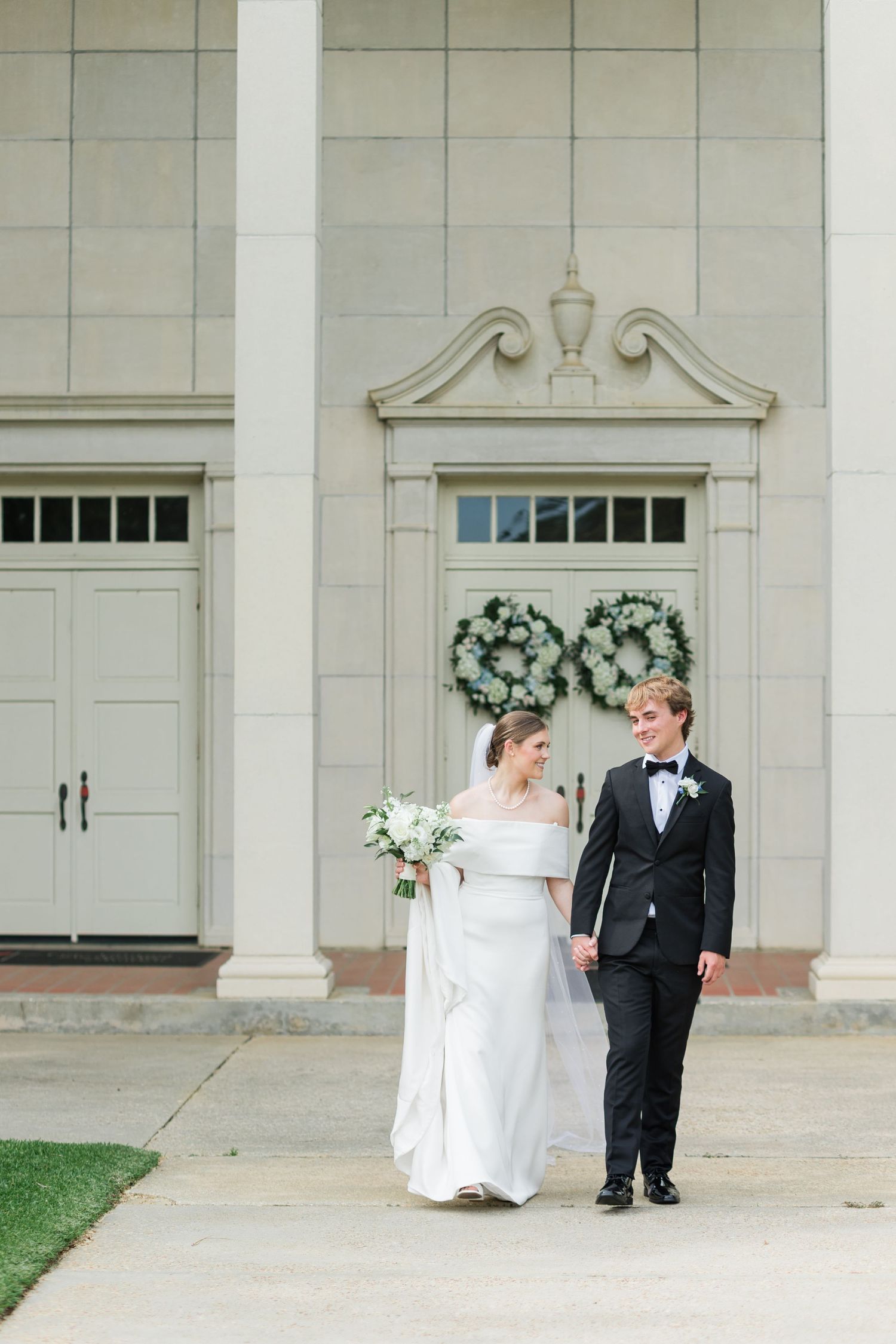 Bride and groom walk hand-in-hand before classic white columned church adorned with greenery wreaths.